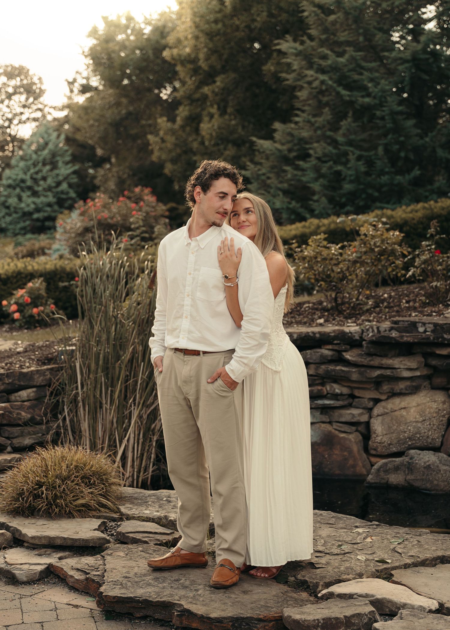 Couple in matching cream outfits posing in a lush garden setting on stone steps at golden hour.