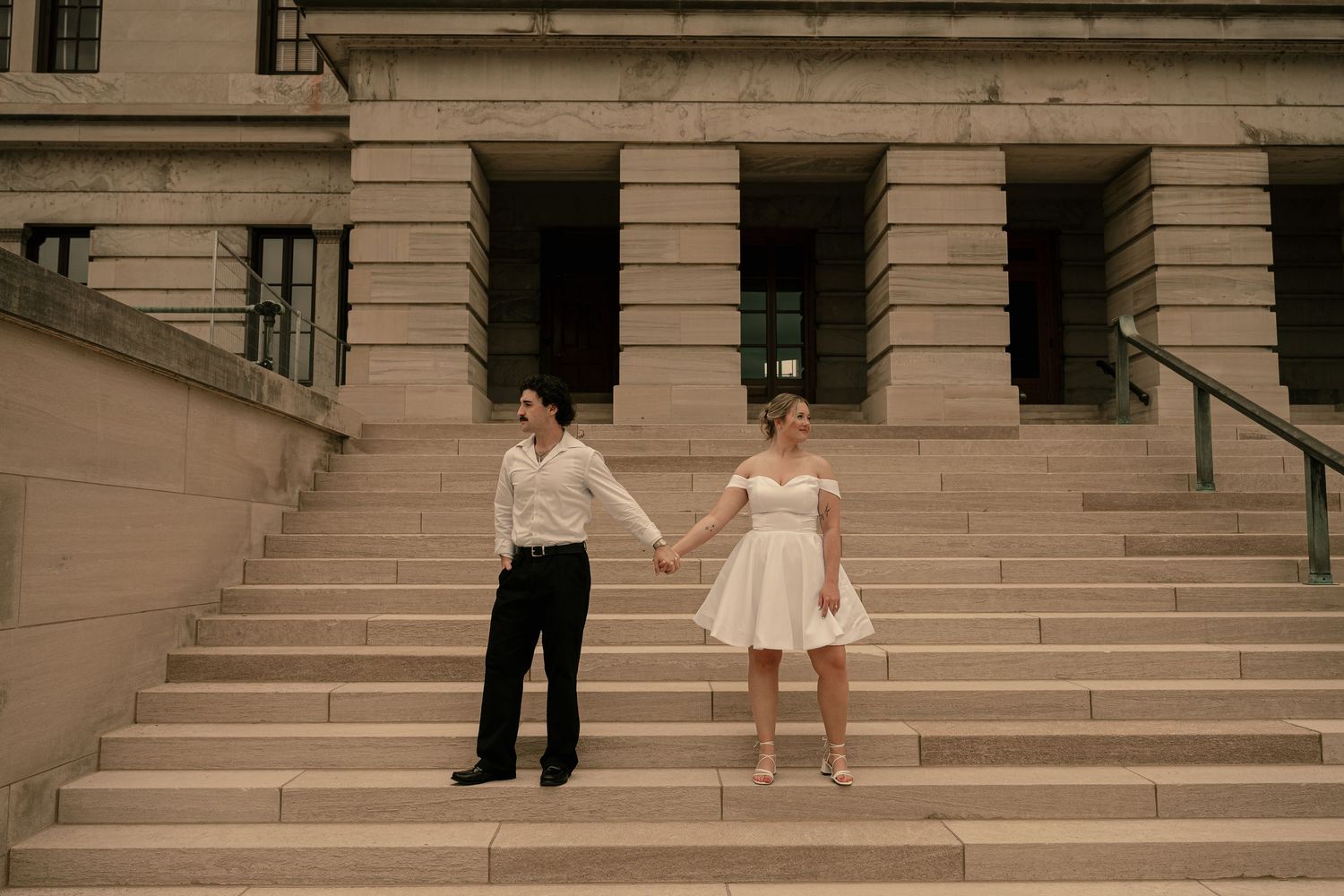 Couple in white outfits holding hands on grand stone steps of a classical building during engagement photo session.