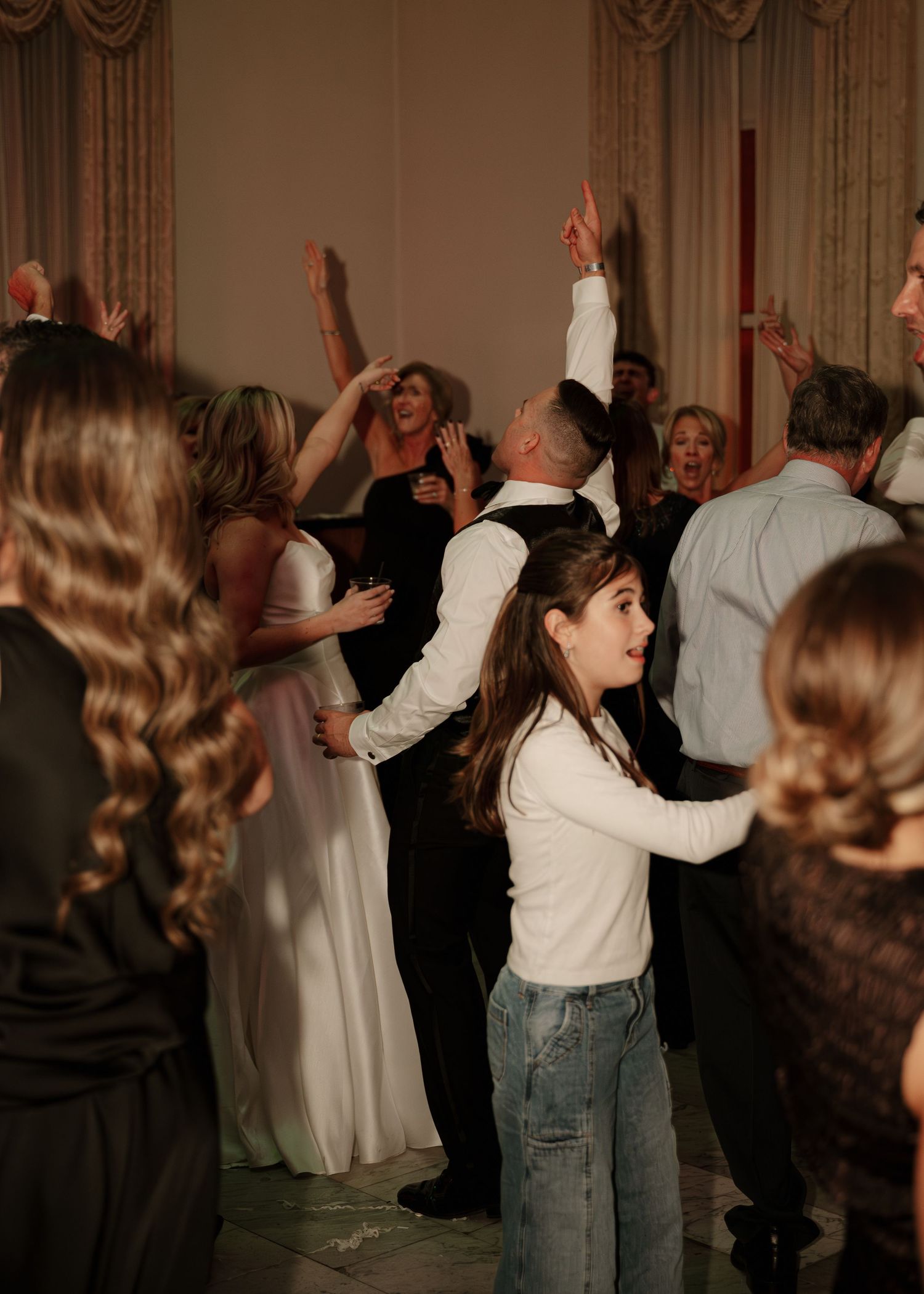 Bride groom and guests celebrate with hands raised on dance floor at lively indoor wedding reception.