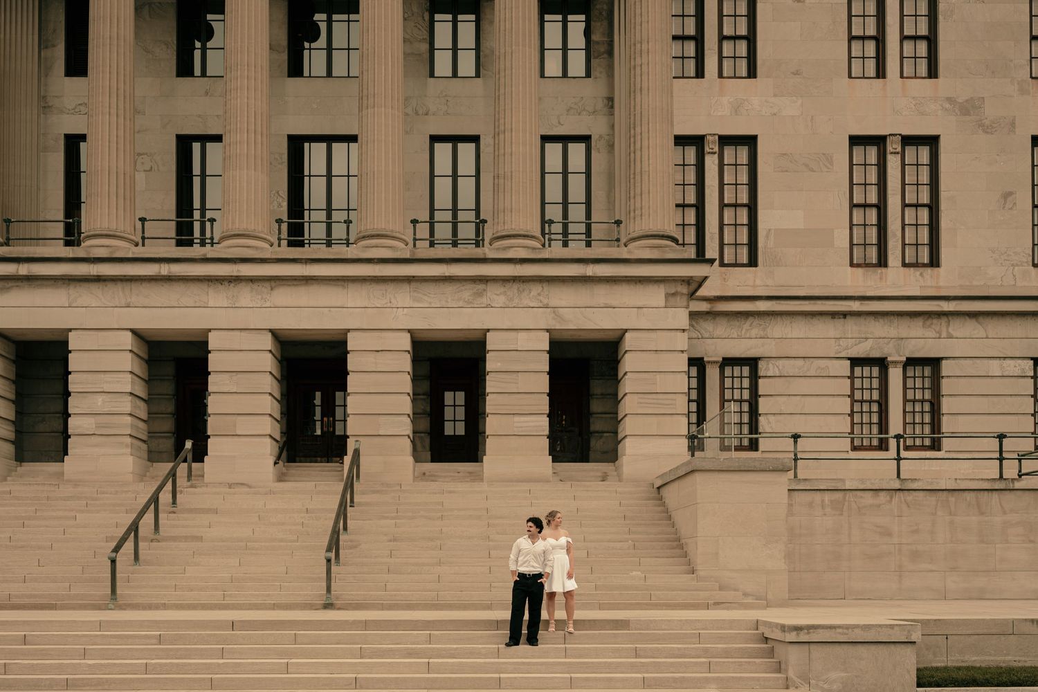 Couple standing on grand stone steps of a neoclassical building with tall columns and large windows.