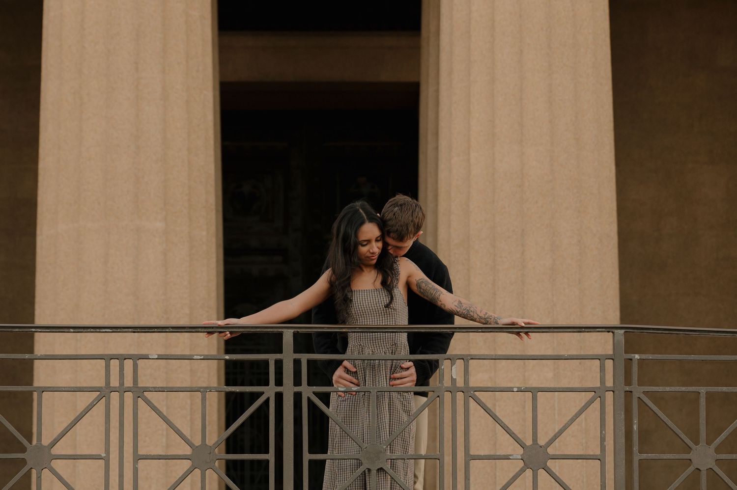 A couple shares an intimate embrace on a balcony between classical columns of a Greek-style building.