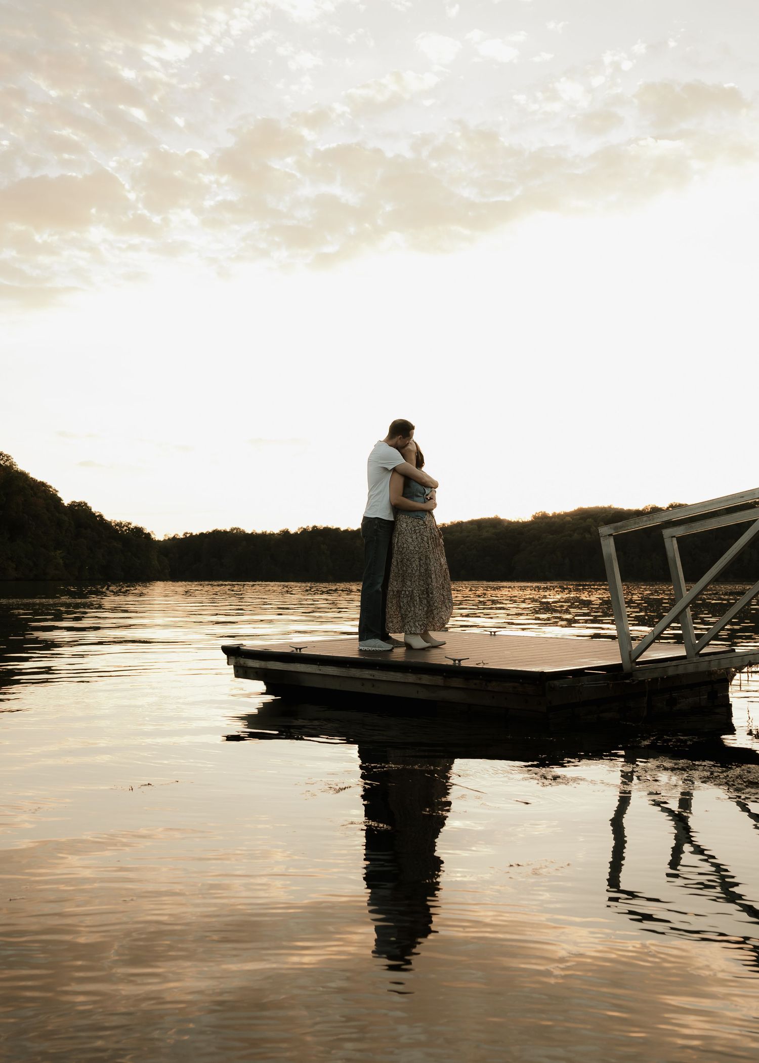 Couple embracing on a wooden dock at sunset over a calm reflective lake.