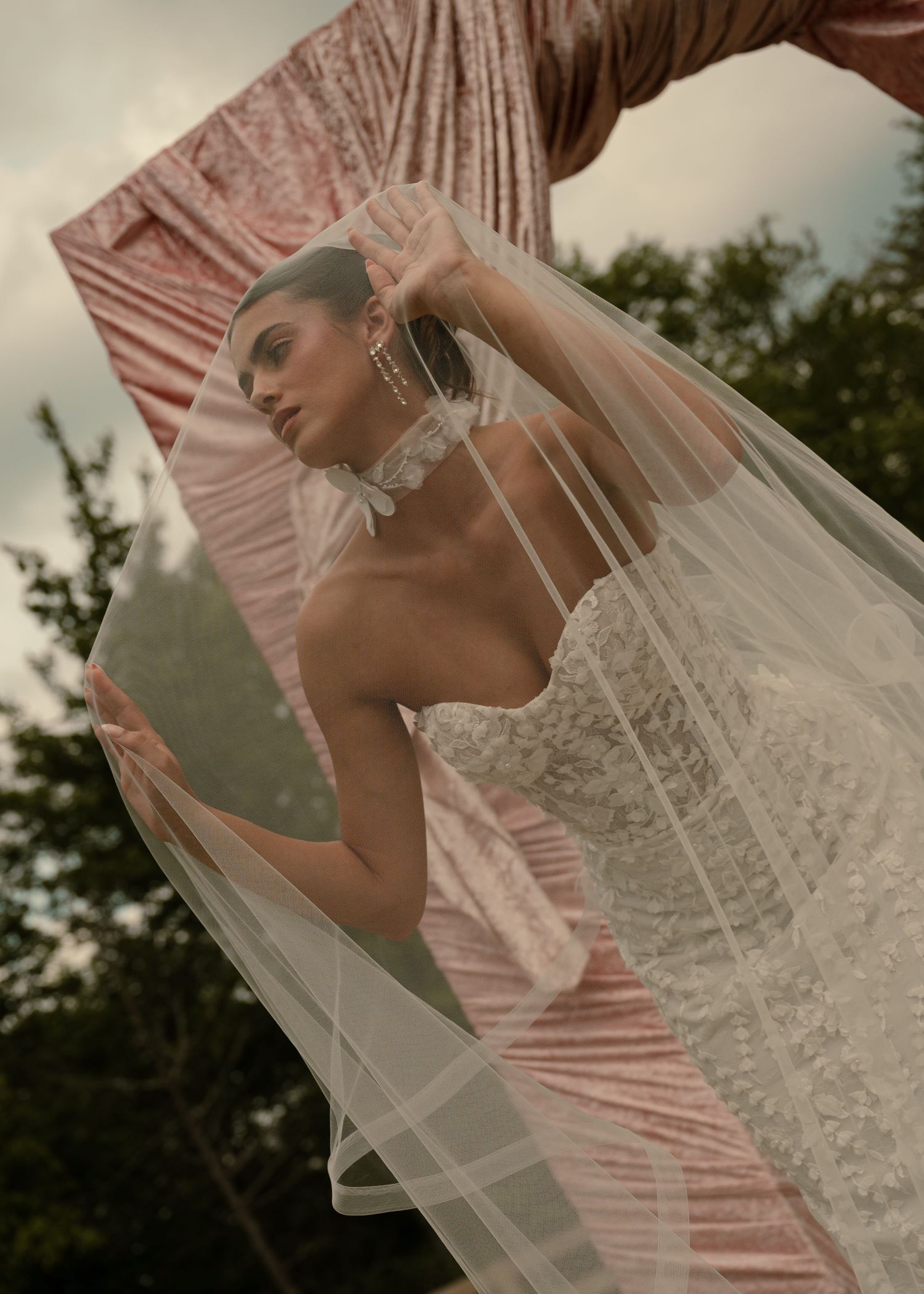 Bride in strapless lace wedding gown and flowing veil poses elegantly beside a pink velvet mirror outdoors.