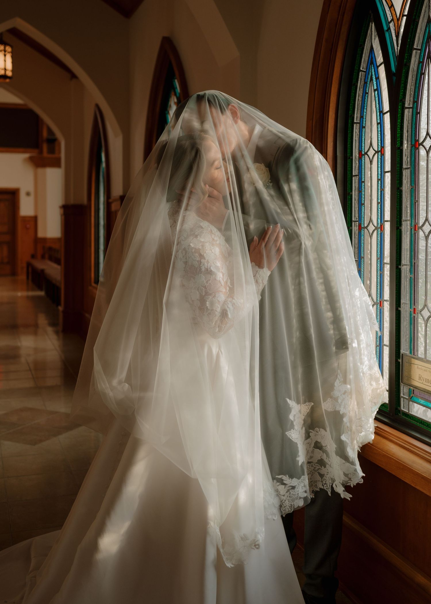 Bride and groom sharing a kiss beneath a lace-trimmed wedding veil in a church with stained glass windows.