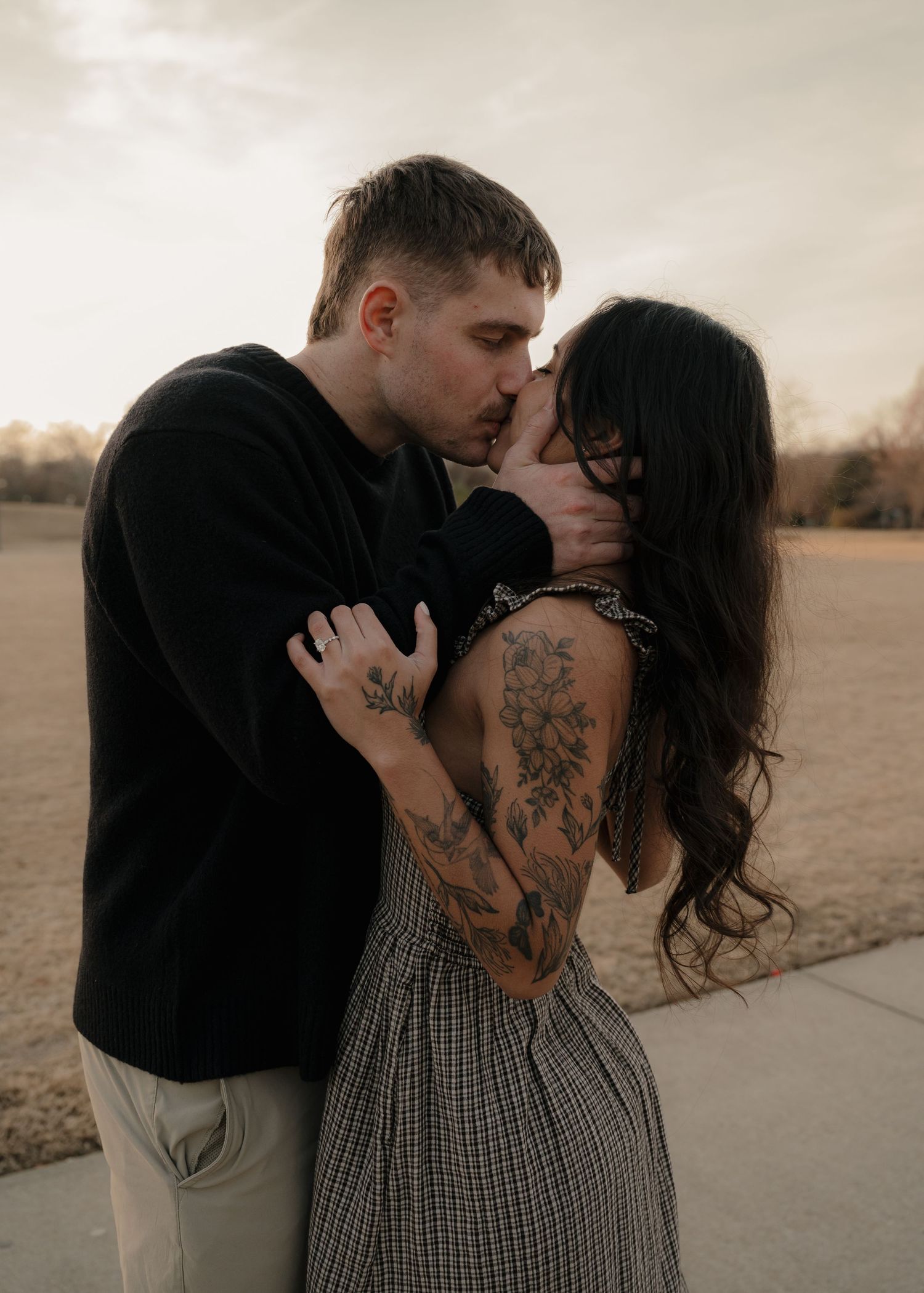 Couple sharing an intimate kiss outdoors at sunset, woman with floral tattoos embraced by man in black sweater.