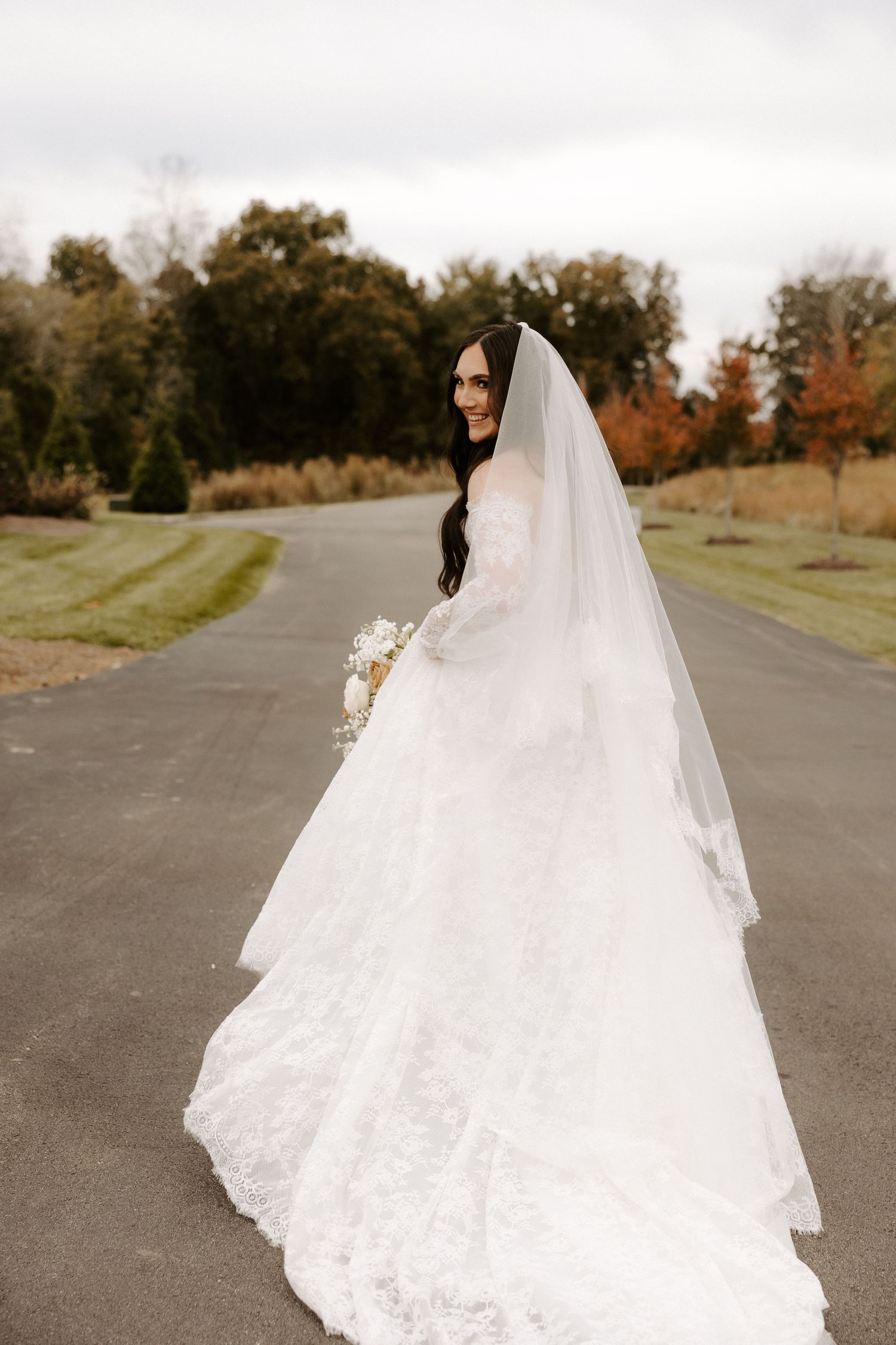 A bride in a long lace wedding gown and veil walks along an outdoor pathway.