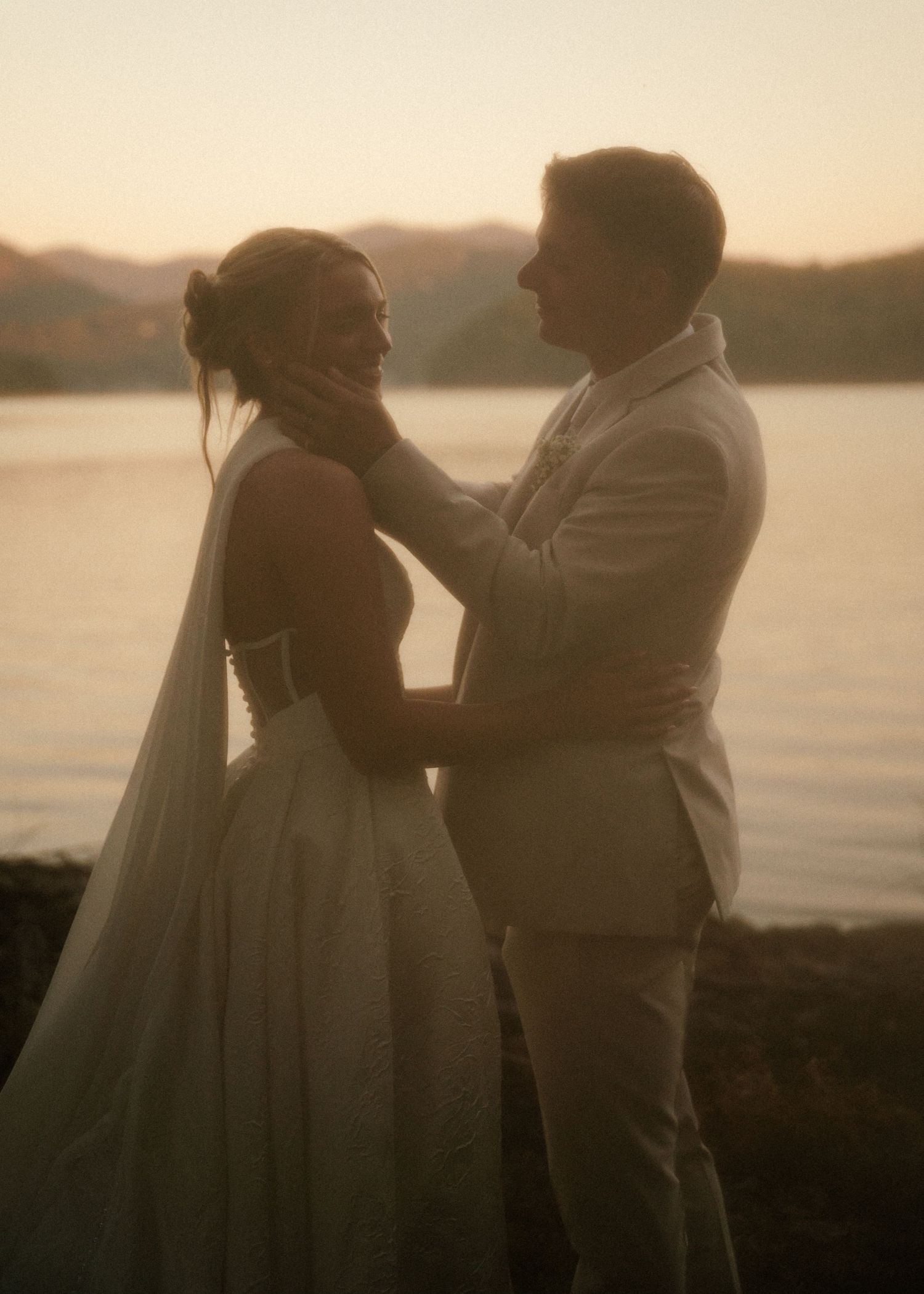 A couple shares a romantic moment at sunset by a lake, the groom tenderly holding the bride's face in his hands.