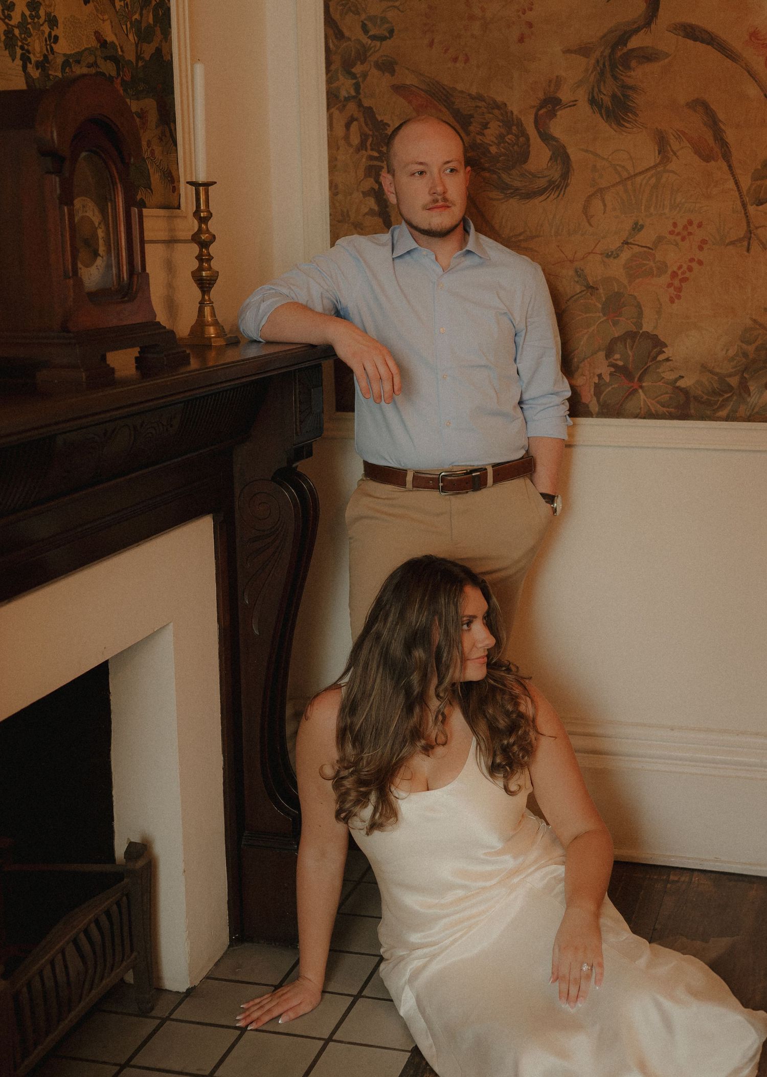 Groom leaning on fireplace mantel while bride sits gracefully on floor in elegant white dress.
