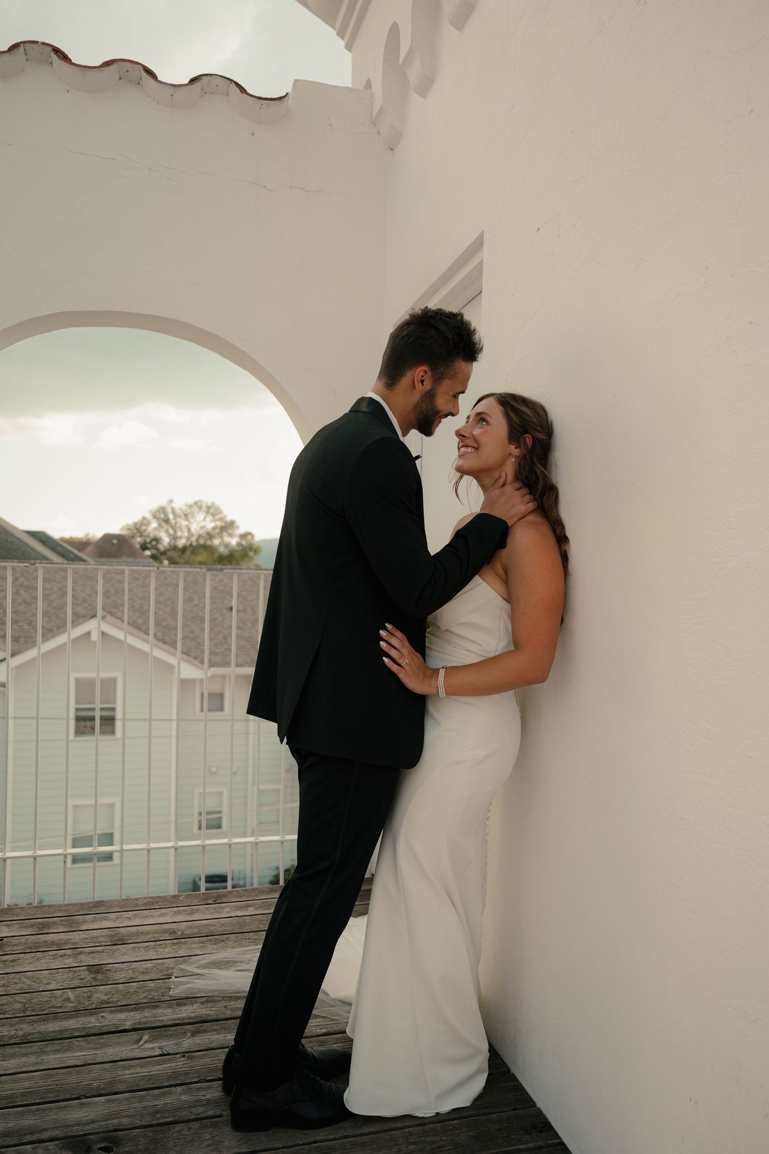 Bride and groom sharing intimate moment leaning against white wall on rooftop balcony at sunset.