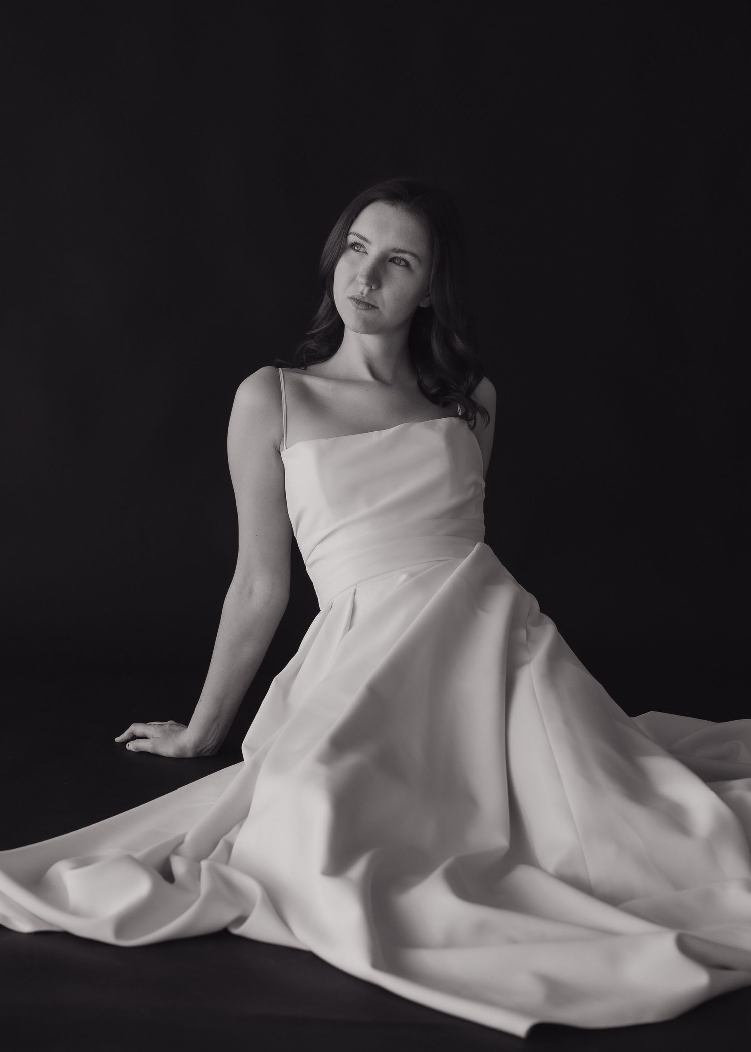 Elegant woman in white satin gown seated gracefully against dark background in black and white.