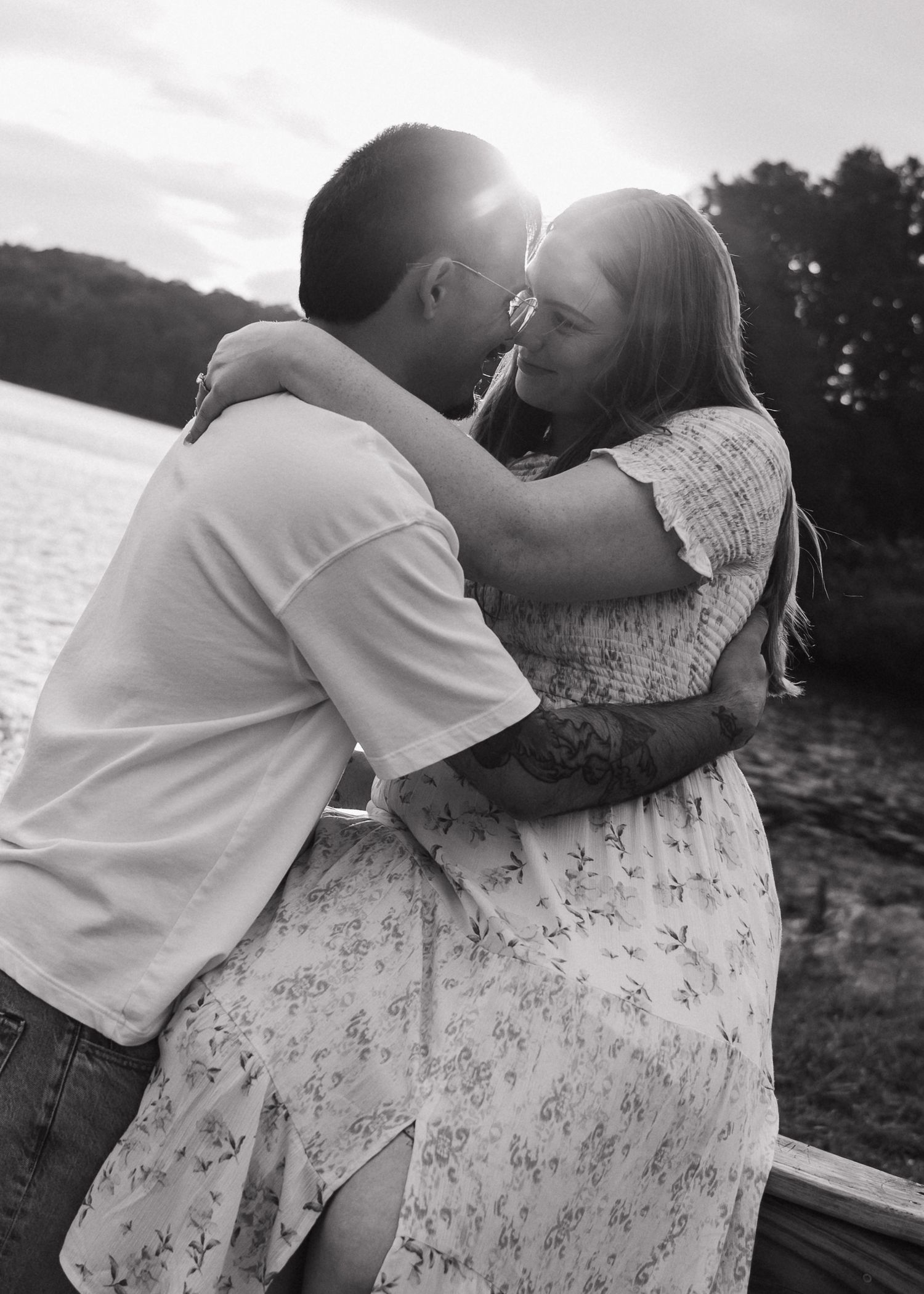 Couple sharing a romantic kiss by a lake at sunset in a black and white photo.