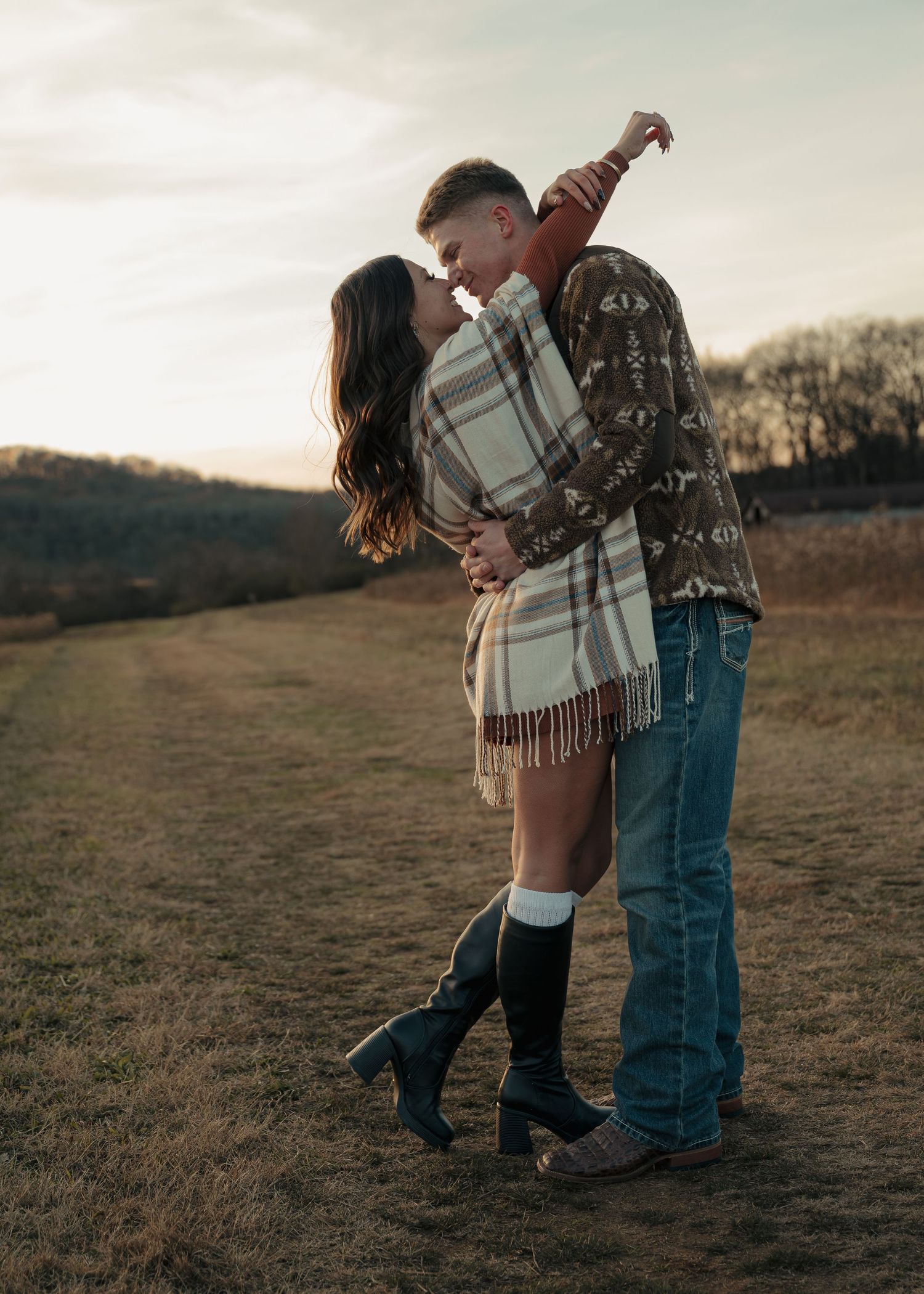 Couple sharing romantic kiss at sunset in open field, woman wrapped in plaid blanket wearing knee-high boots.