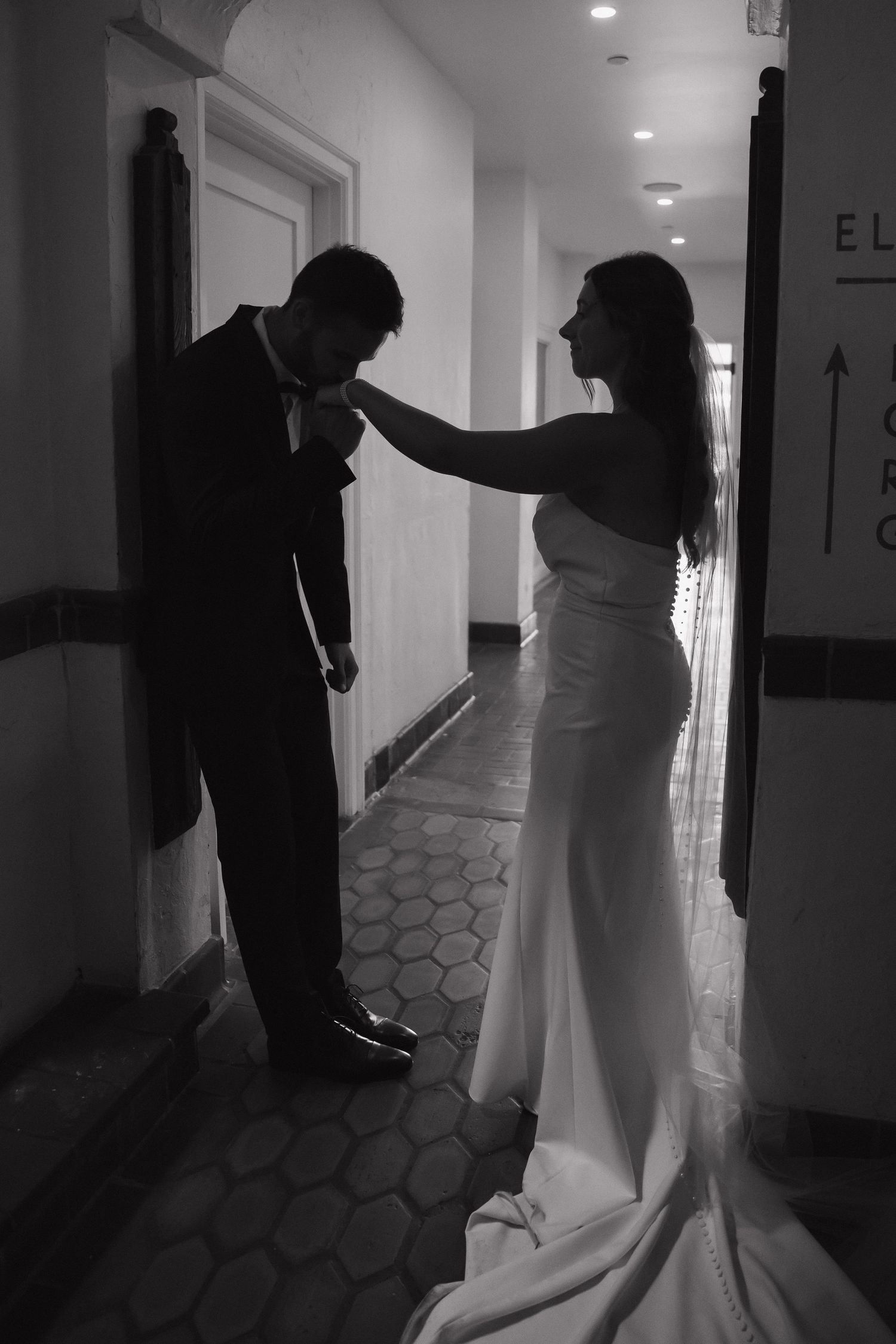 Black and white photo of bride adjusting groom's boutonniere in elegant hotel hallway.