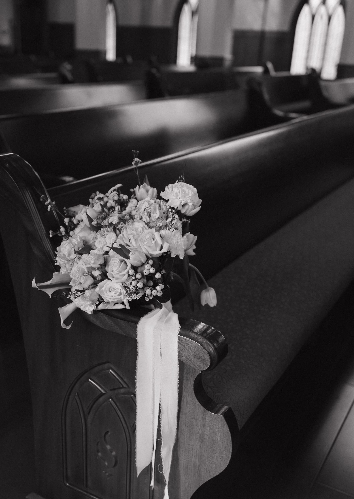 Black and white photo of a floral wedding bouquet resting on a wooden church pew.