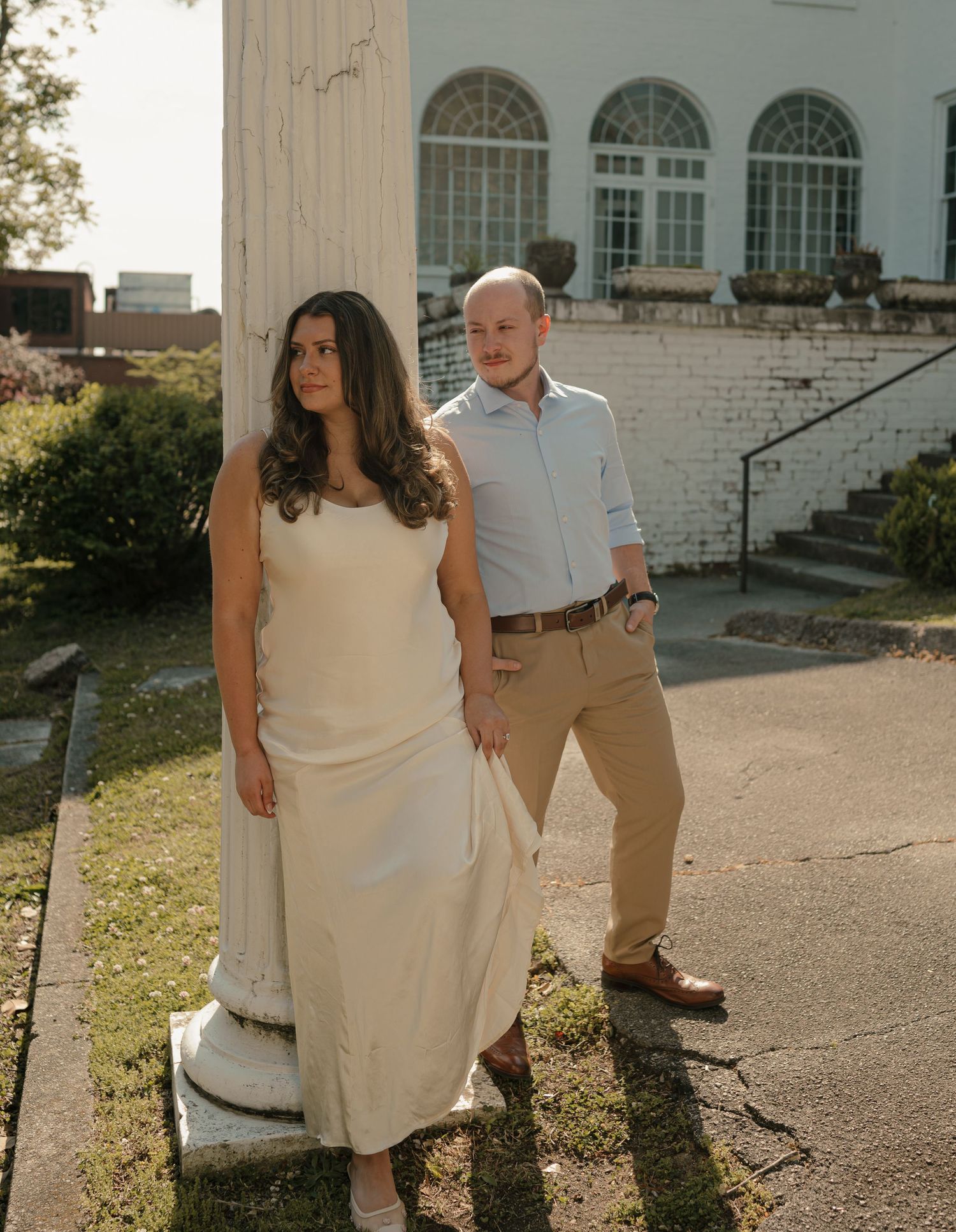 Elegant couple posing by white column outside historic building, woman in cream dress, man in khakis.