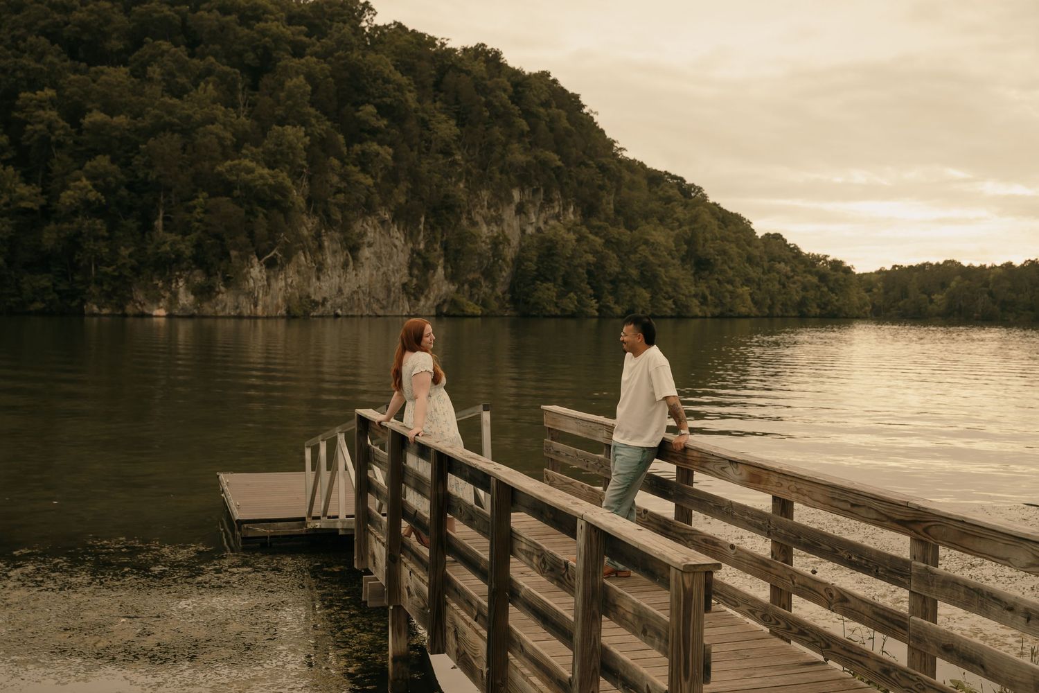 Couple standing on a wooden dock overlooking a serene lake surrounded by lush green cliffs at dusk.