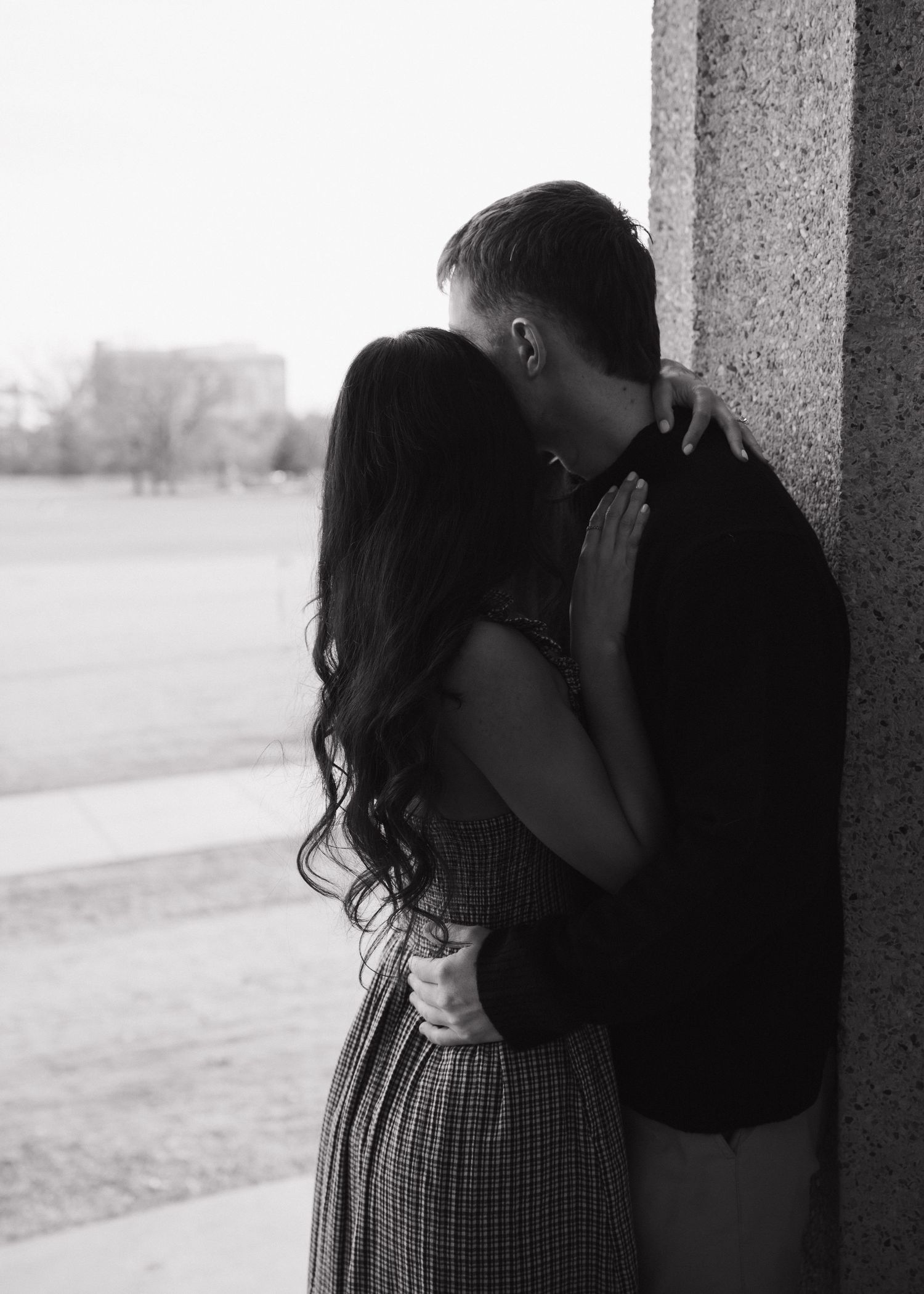 Black and white photo of couple kissing against stone column outdoors.