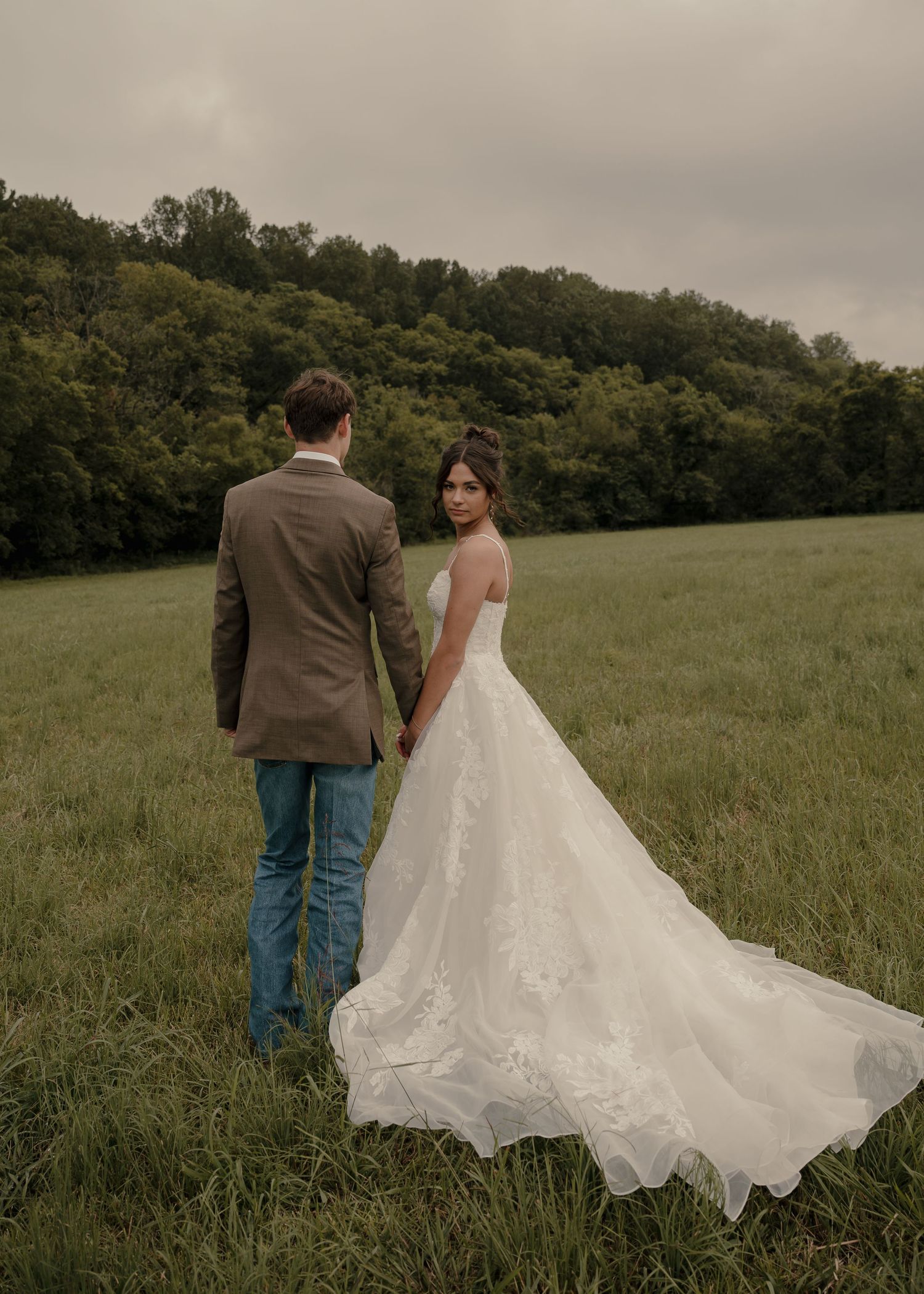 Bride in lace gown holding groom's hand in a green countryside field.