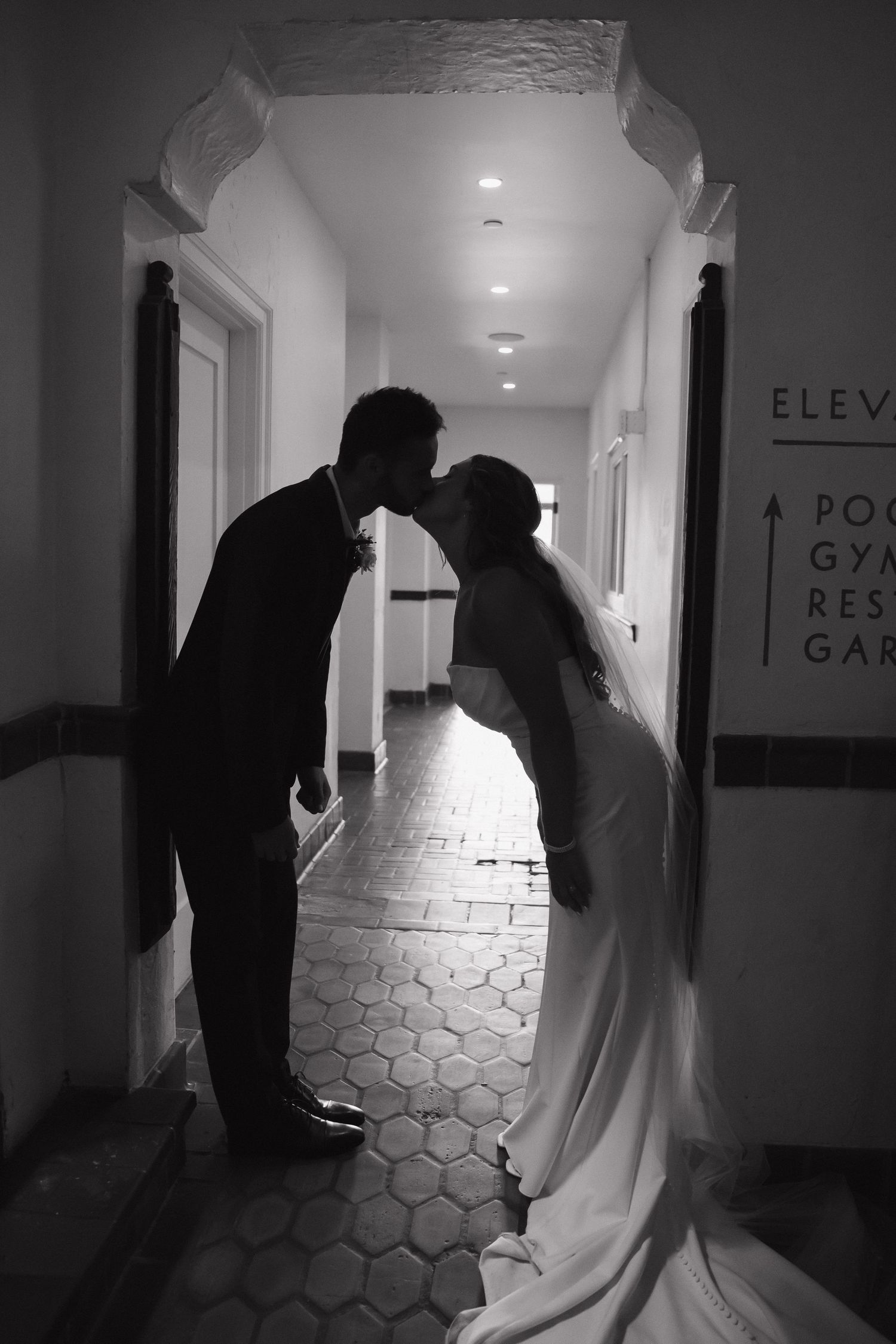 Black and white silhouette of bride and groom sharing a kiss in hotel hallway on wedding day.
