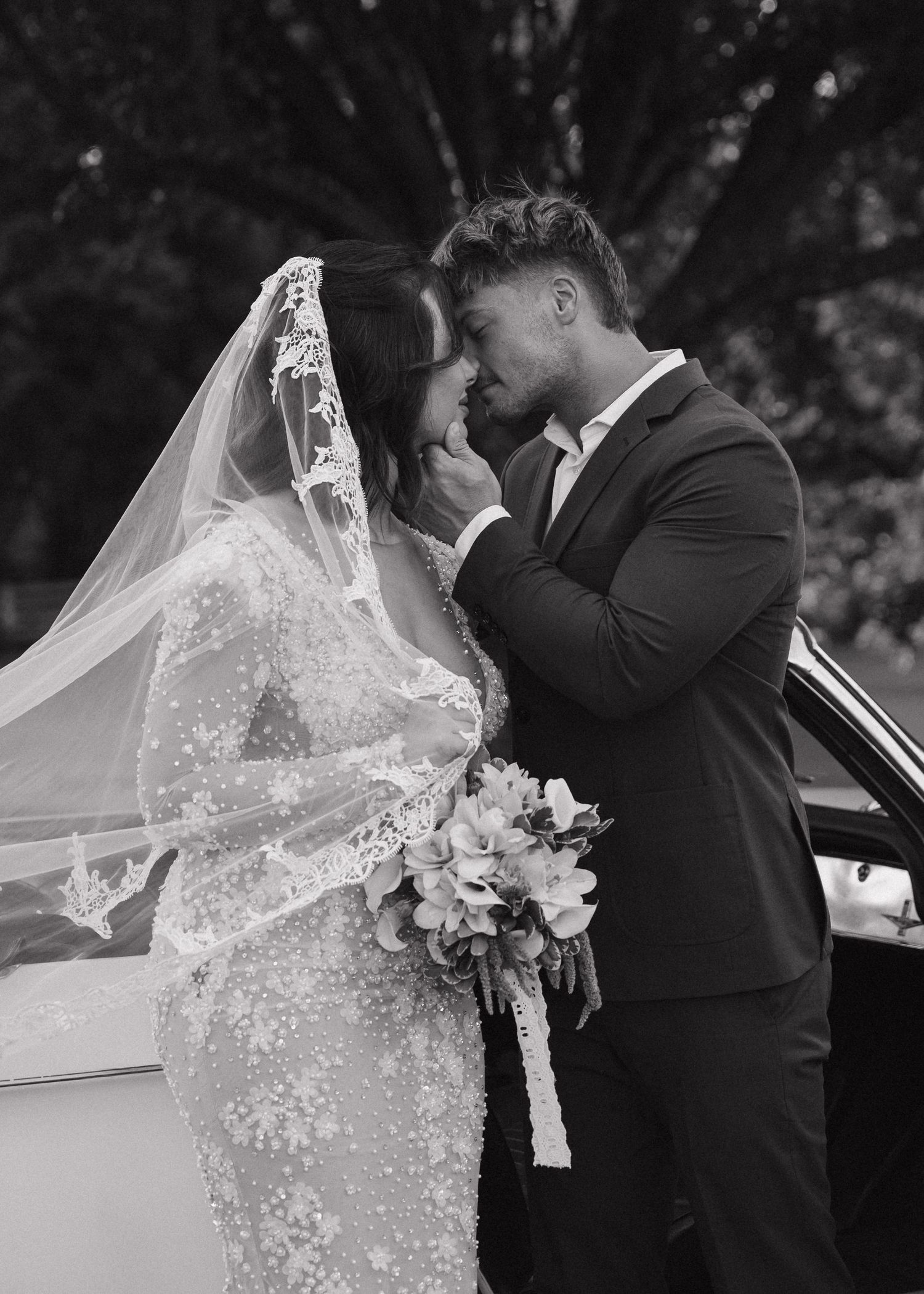 Bride in lace veil and groom in black suit share intimate moment by vintage car, black and white.