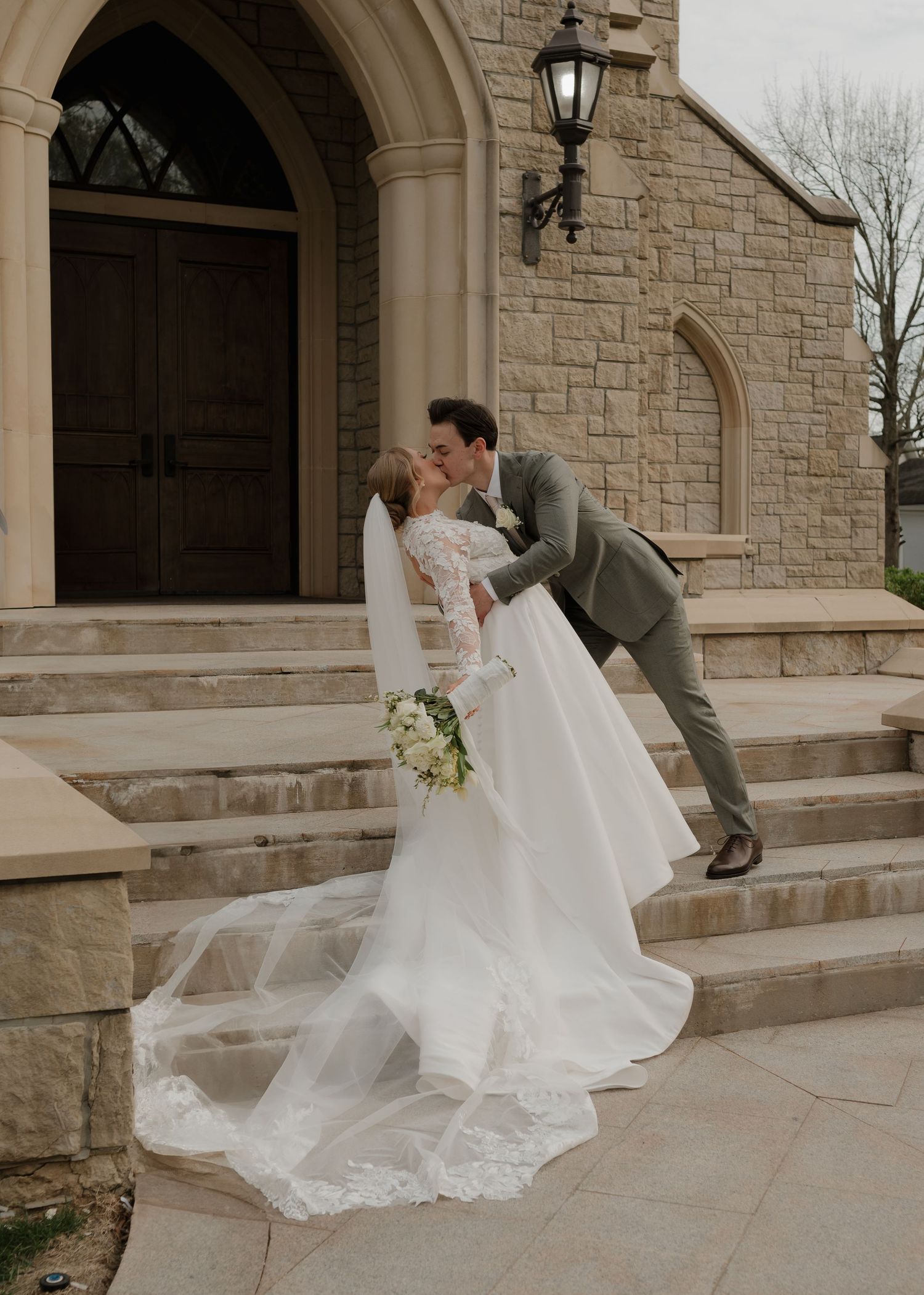 Groom dipping bride for a kiss on the steps of an elegant stone church on wedding day.