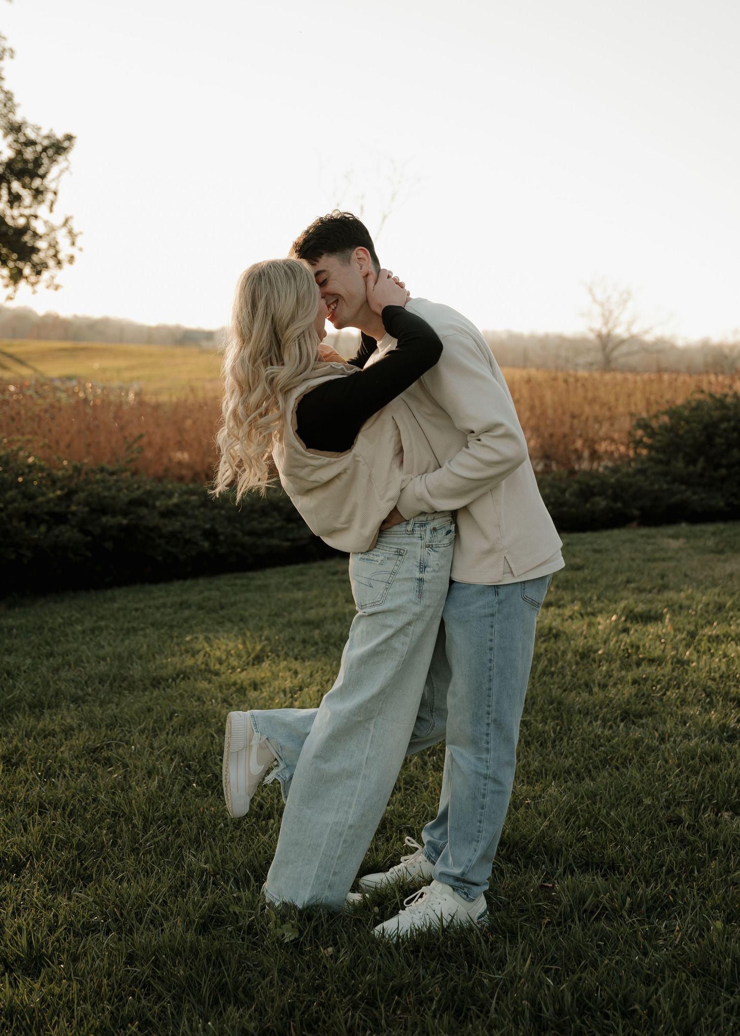 A couple shares a passionate kiss while embracing on a green lawn at golden hour with autumn foliage behind them.