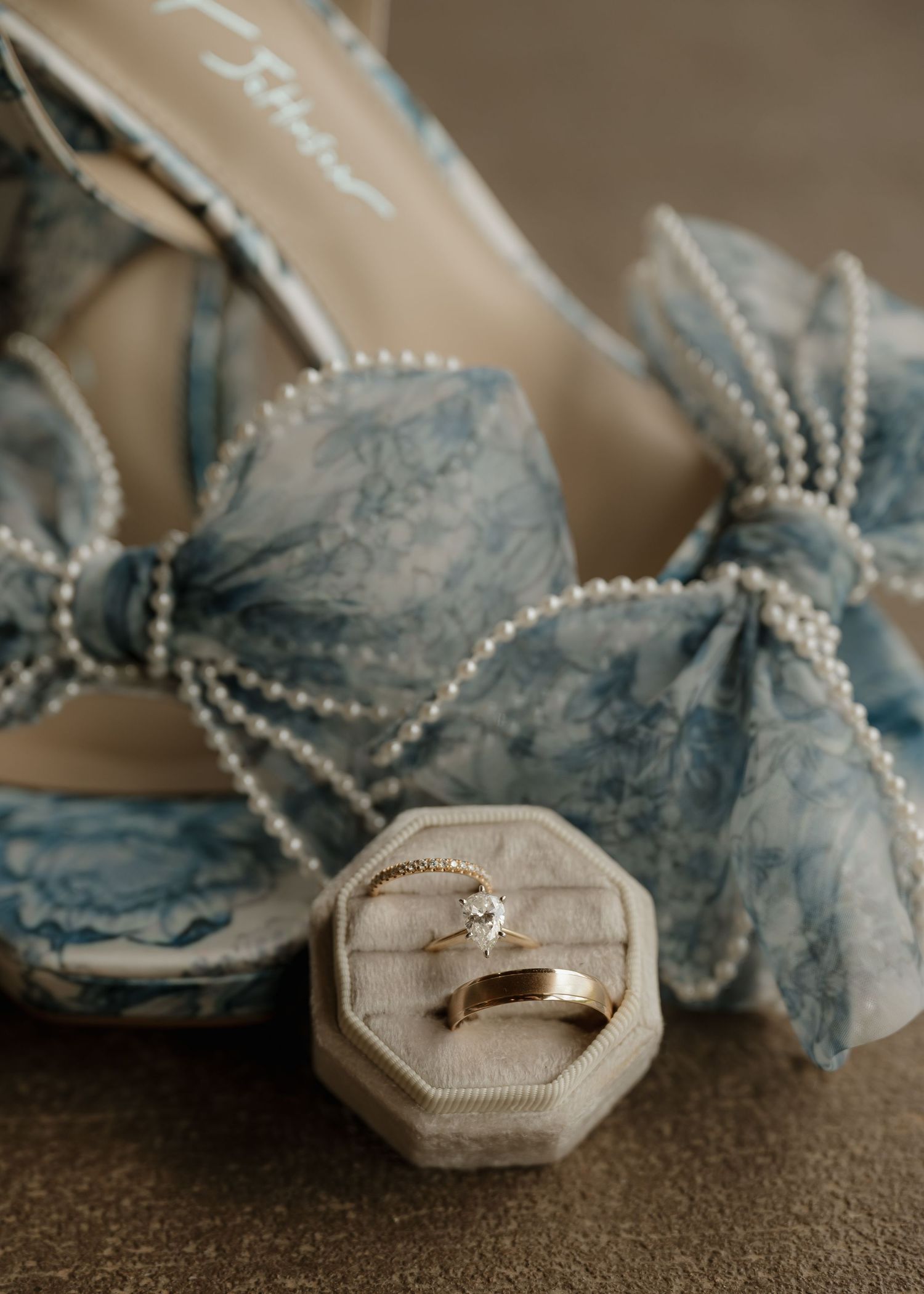 Blue floral bridal heels with pearl bow detail beside gold engagement ring and band in velvet ring box.