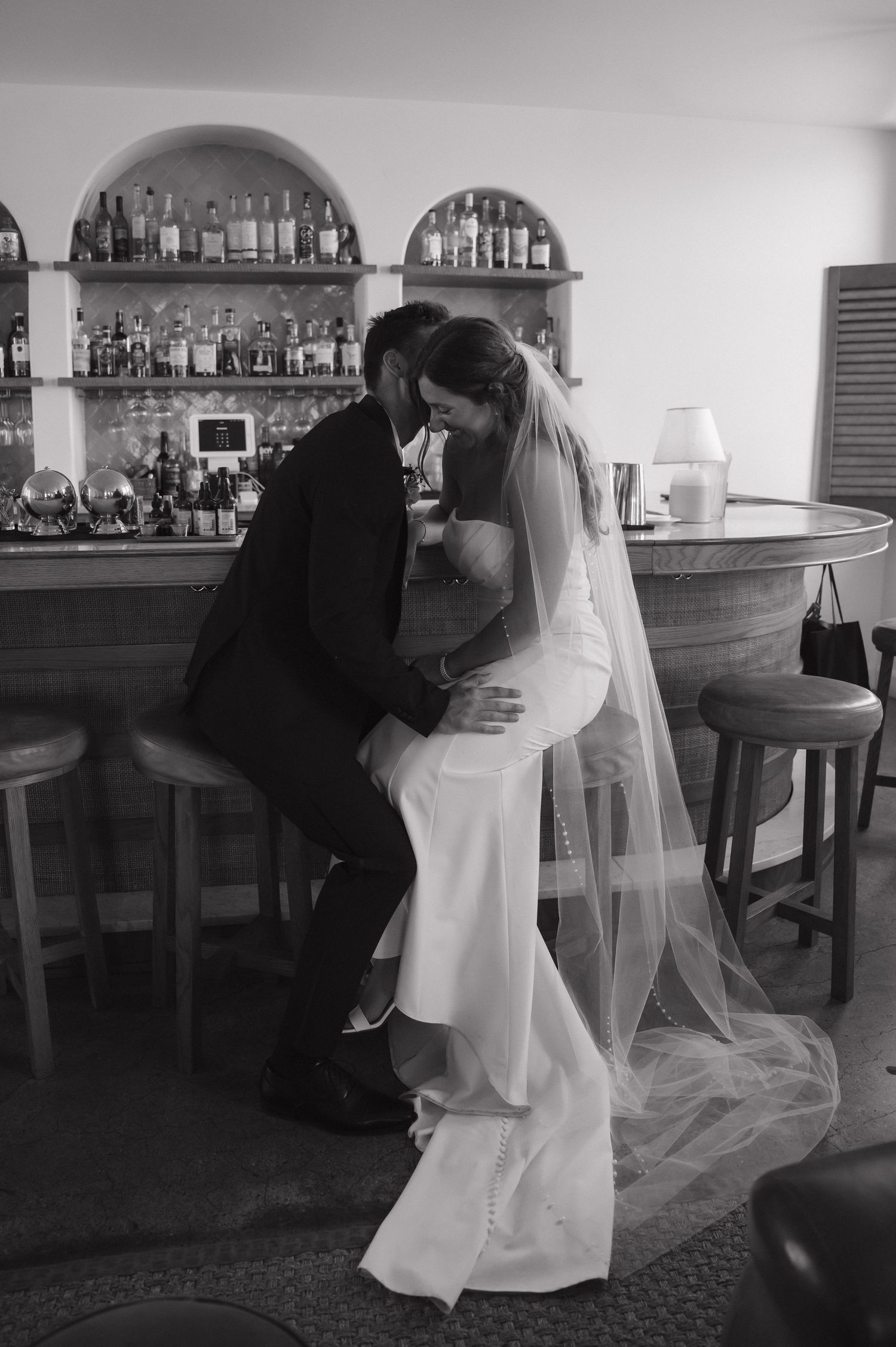 Black and white photo of newlyweds kissing at a bar, bride wearing a veil.