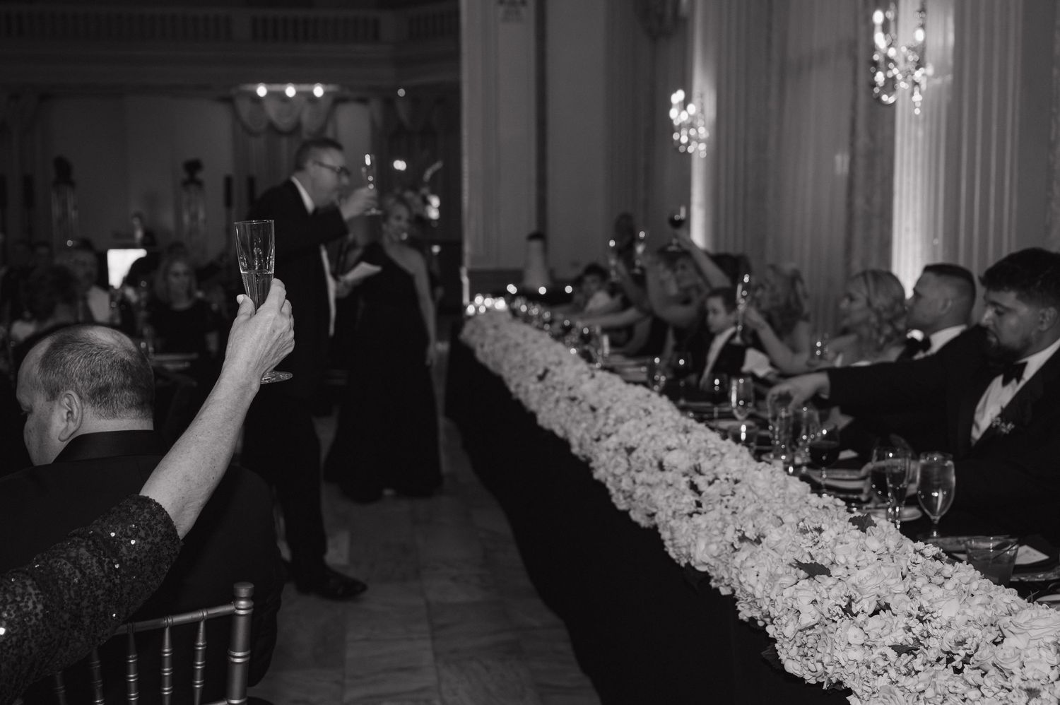 Black and white photo of wedding guests at a long floral-decorated head table during a formal reception toast. Downtown Columbus Wedding.