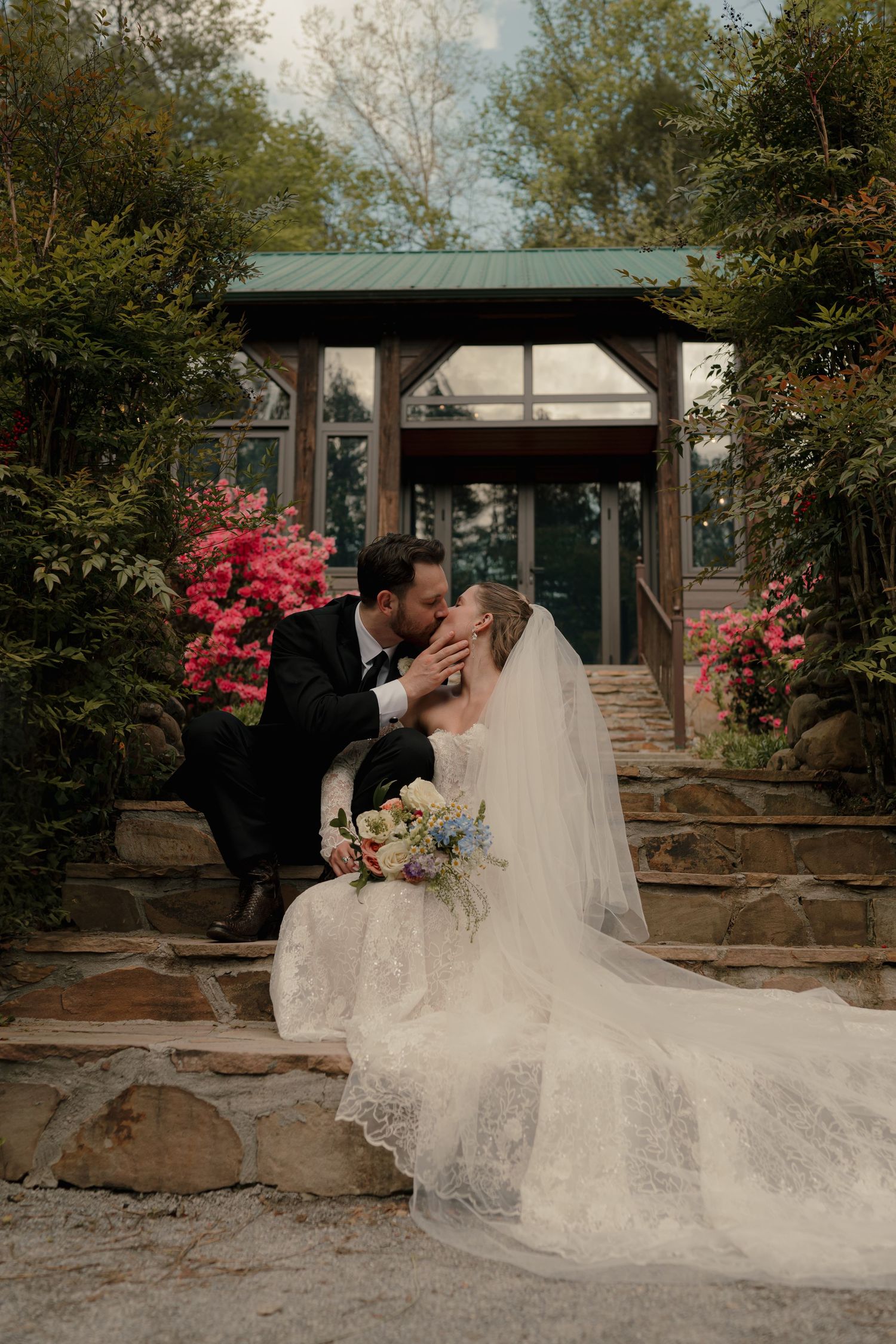 Bride and groom sharing a kiss on stone steps surrounded by blooming pink flowers outdoors.