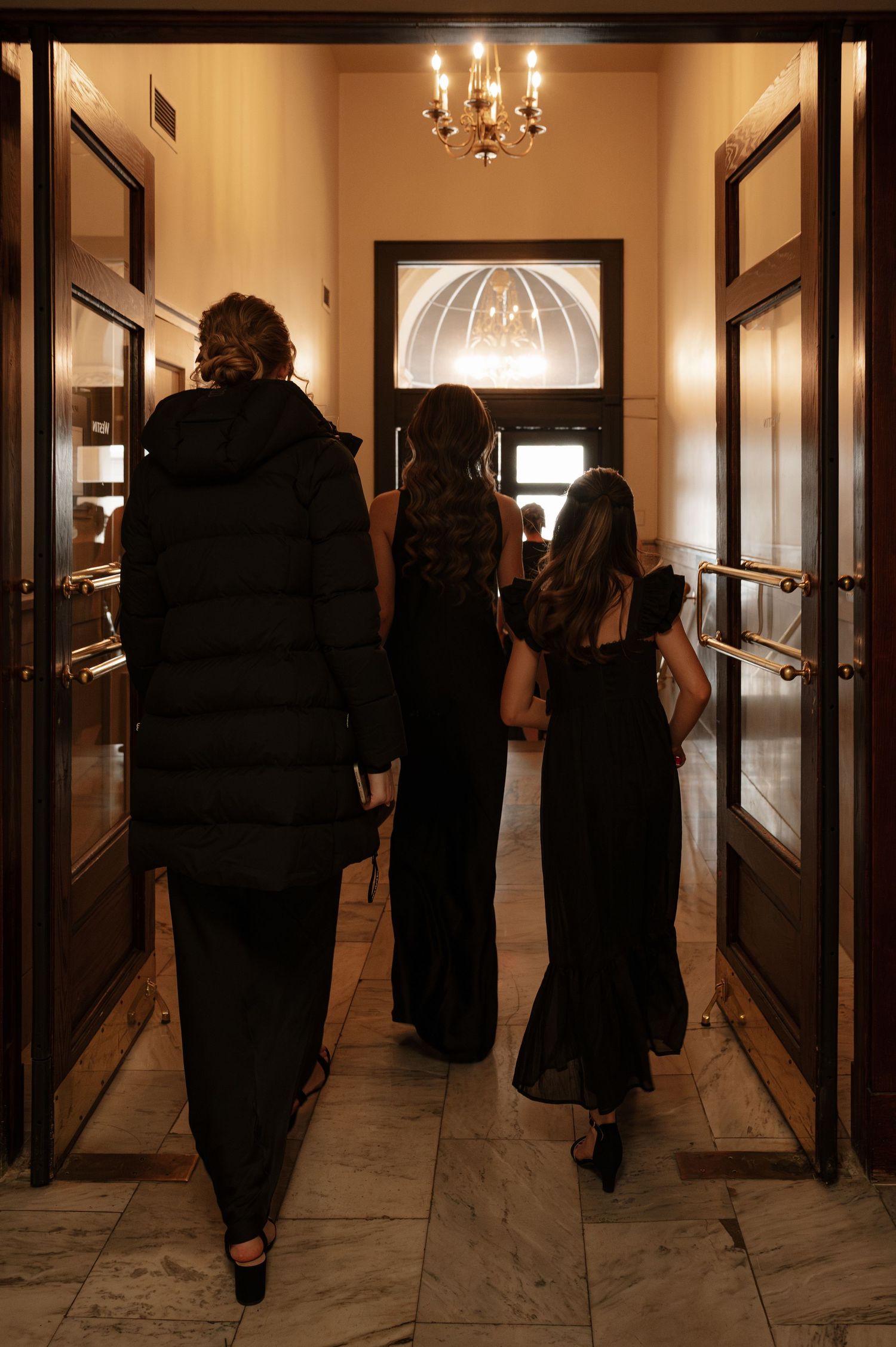Three women in black formal attire walking through ornate venue doors beneath a chandelier. Downtown Columbus Ohio Wedding at The Westin.