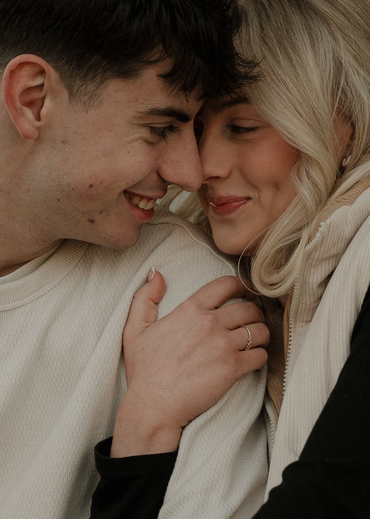 A smiling couple shares a tender forehead-to-forehead moment, the woman clutching the man's cream sweater closely.