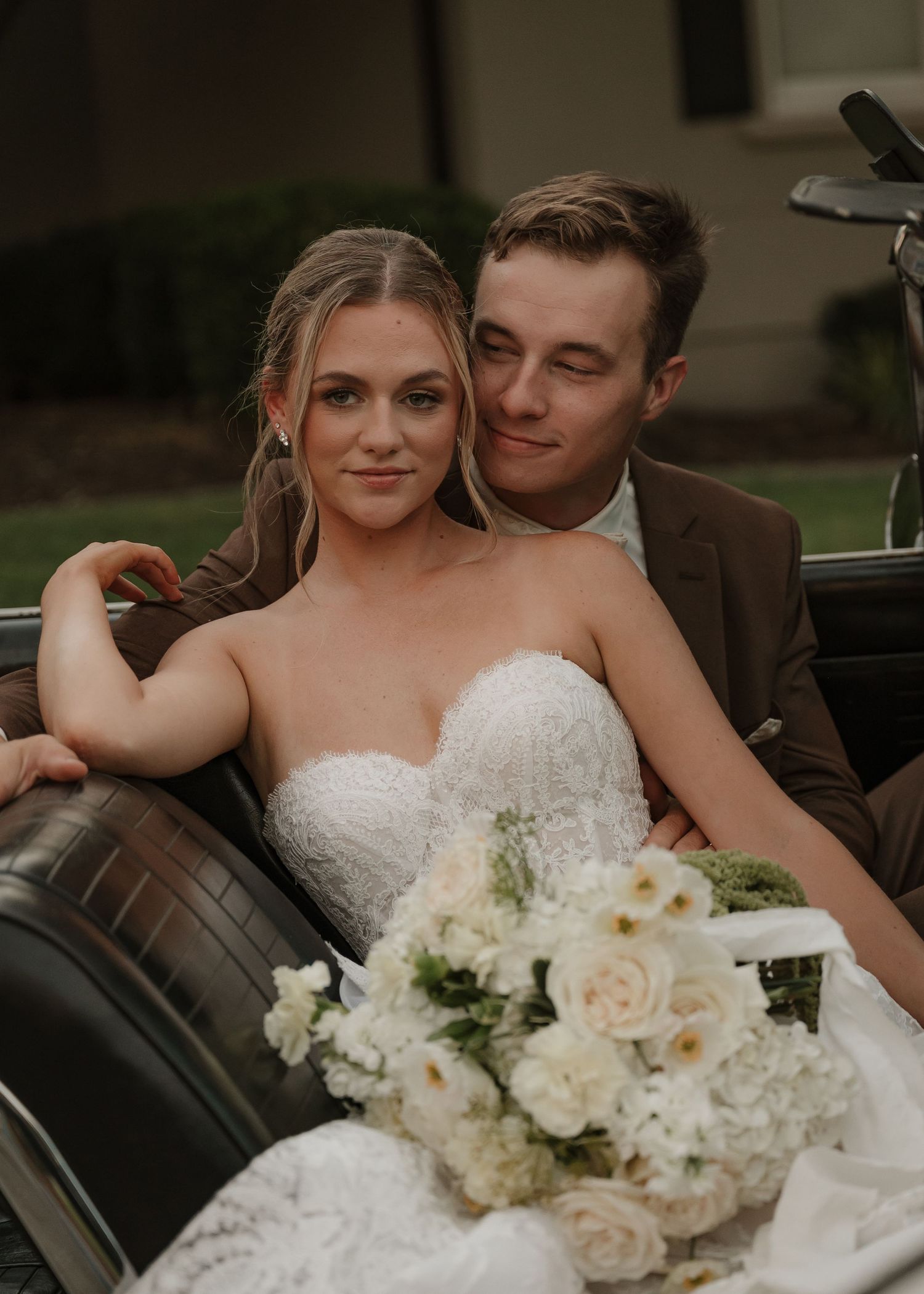 Bride and groom sitting in vintage car with white floral bouquet during wedding portrait session.
