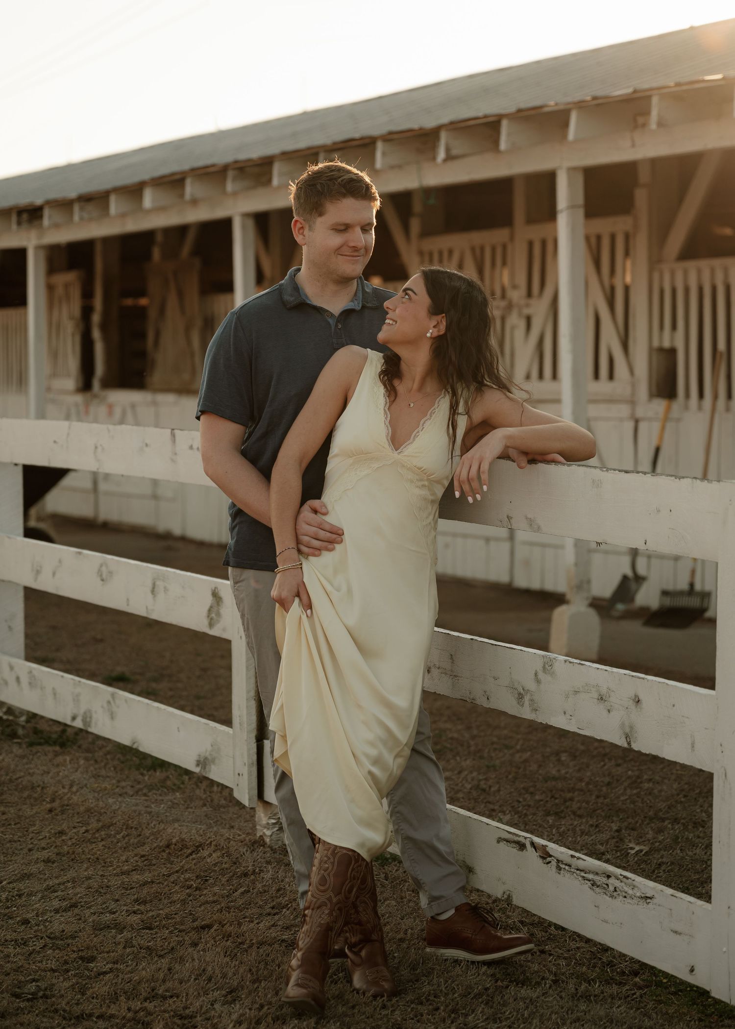 Couple posing together near white fence at sunset with woman in cream dress and cowboy boots.