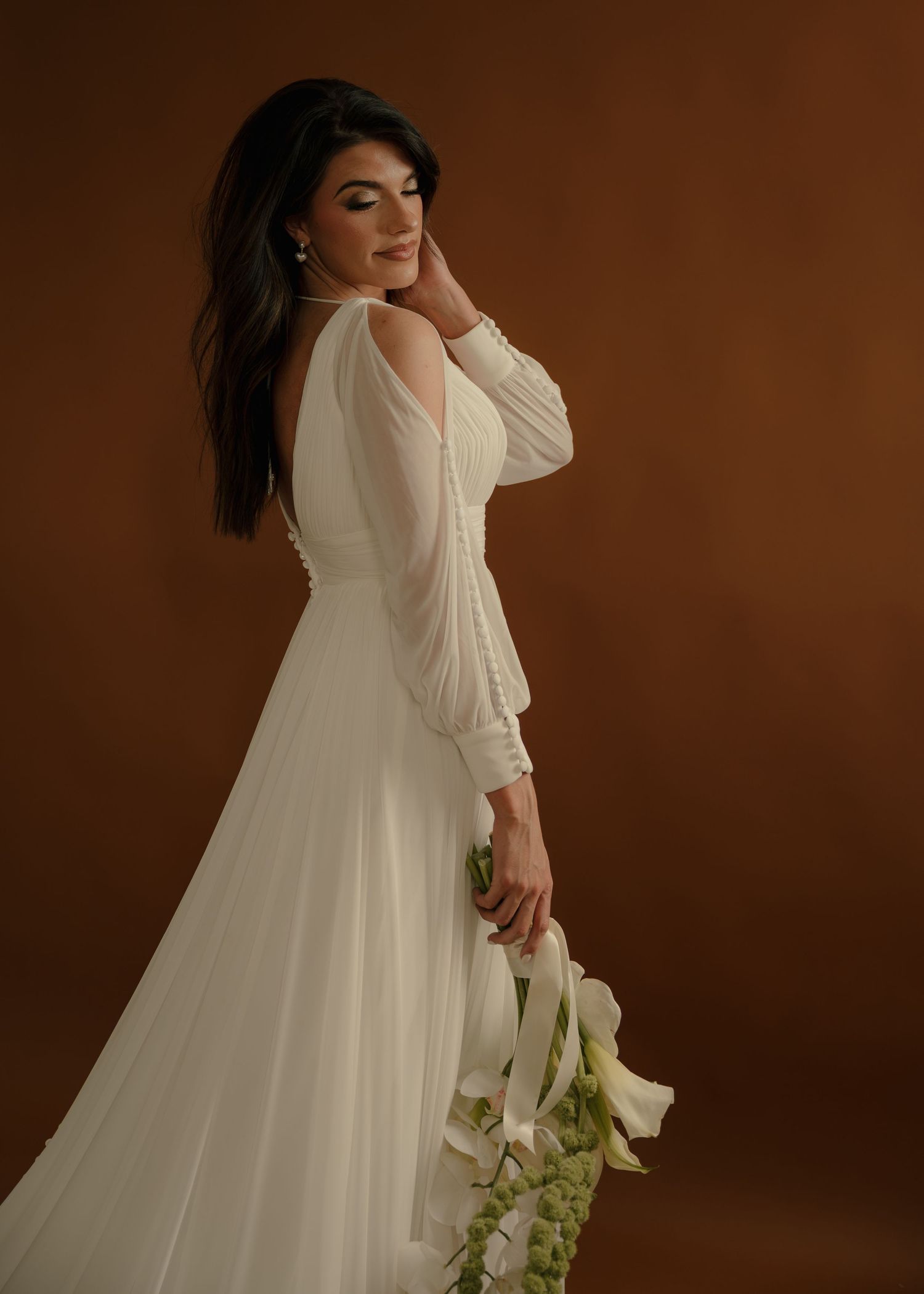 An elegant bride in a flowing white gown holds calla lilies against a warm brown backdrop.
