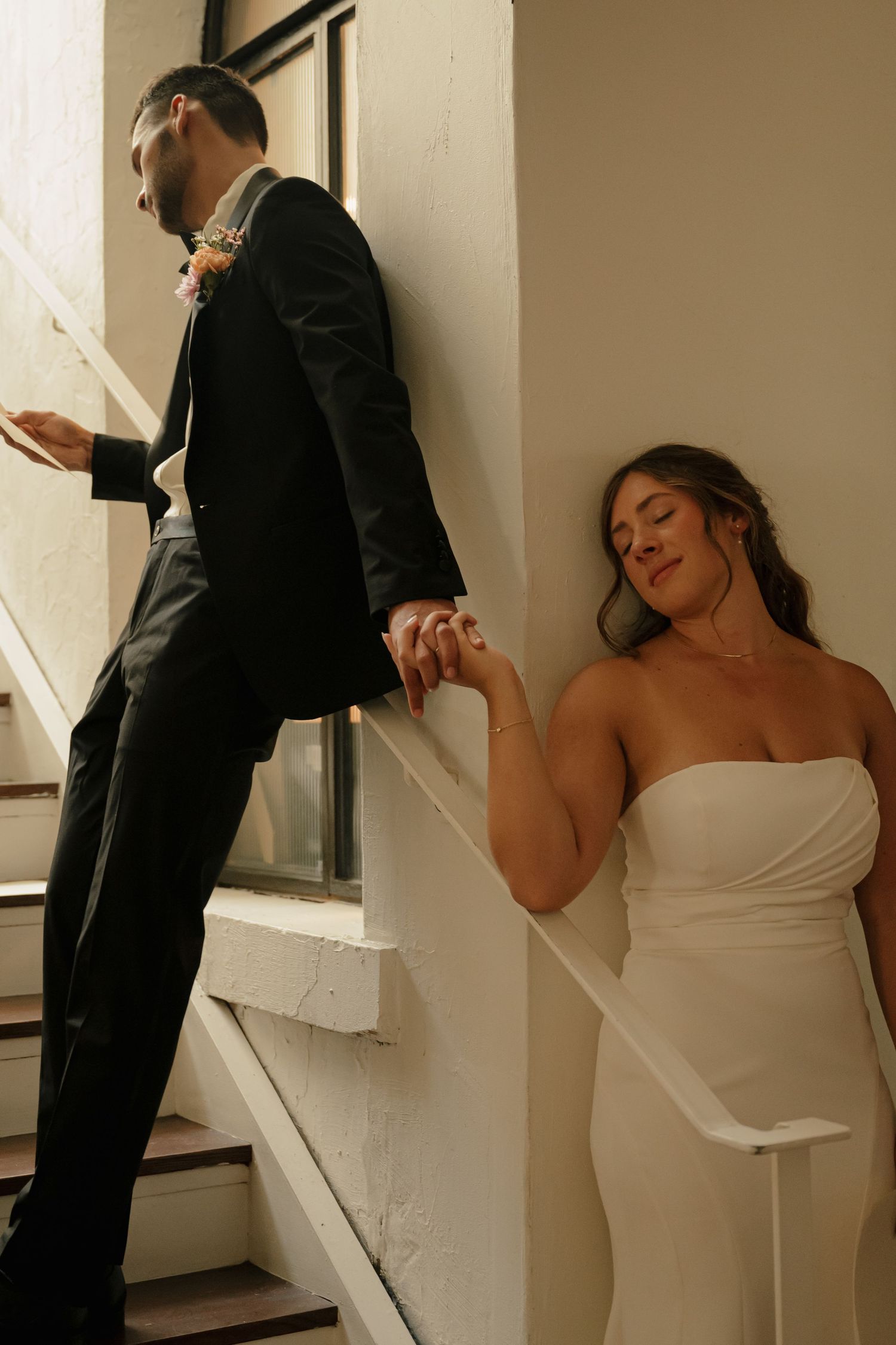 Bride and groom holding hands on a staircase in soft window light.