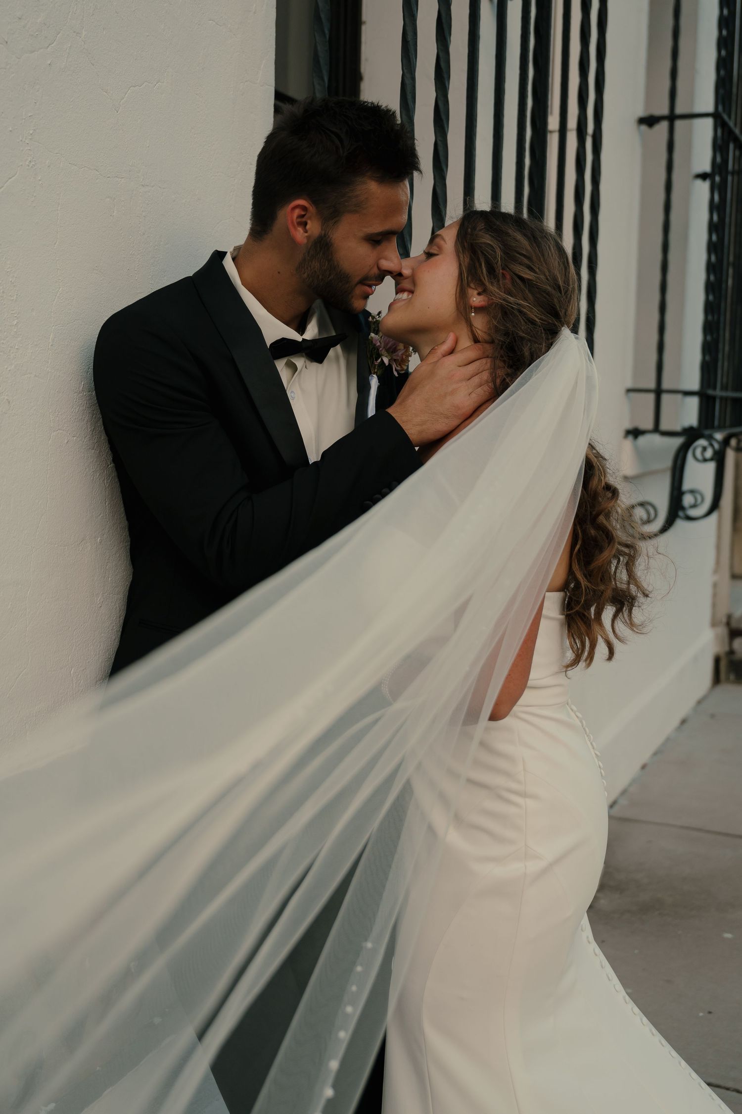 Groom in black tuxedo gently holding bride's face for a kiss as her veil flows in the breeze.