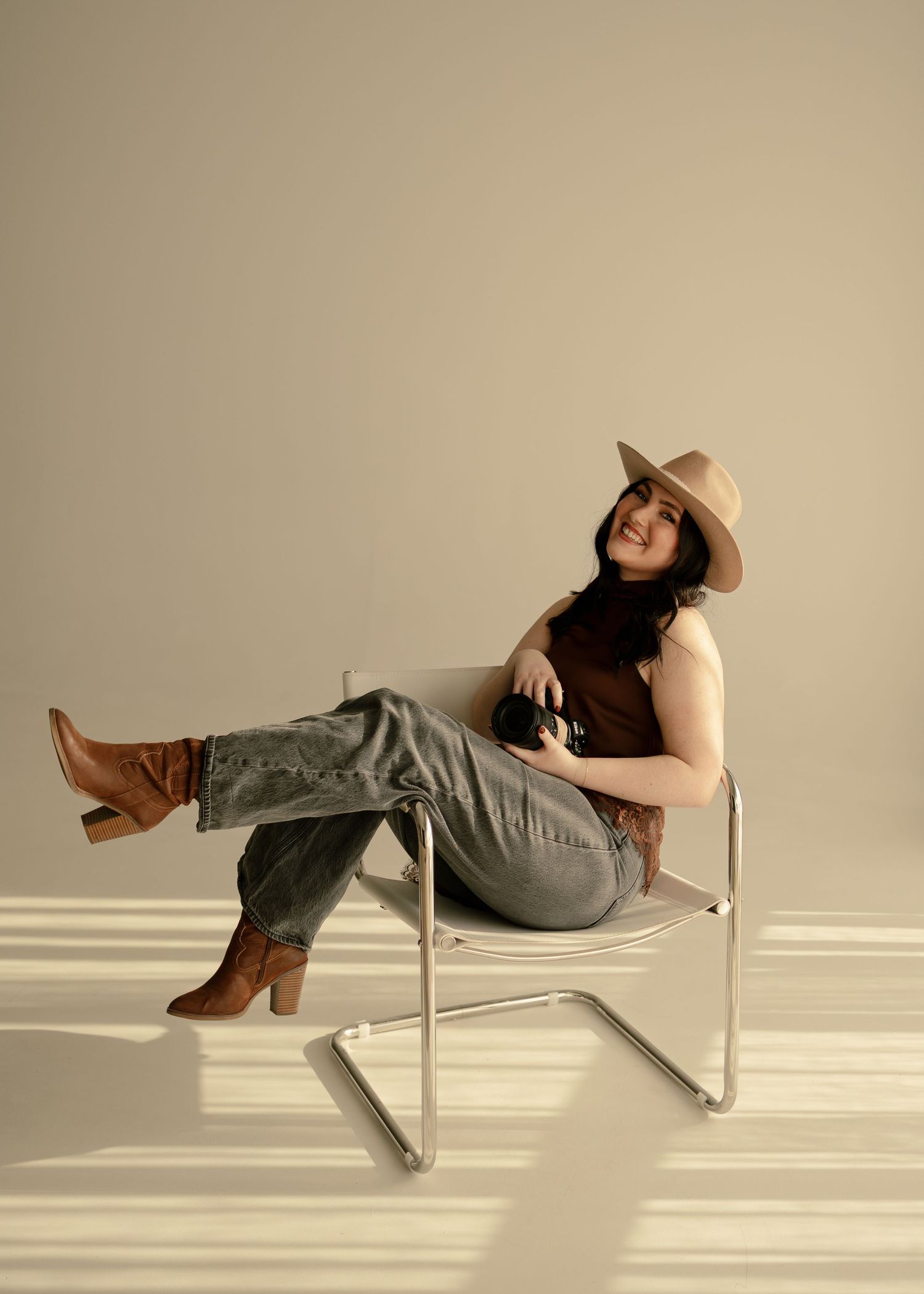 Woman in tan hat, brown top, gray jeans, and brown boots sits relaxed in a modern chair holding a camera.