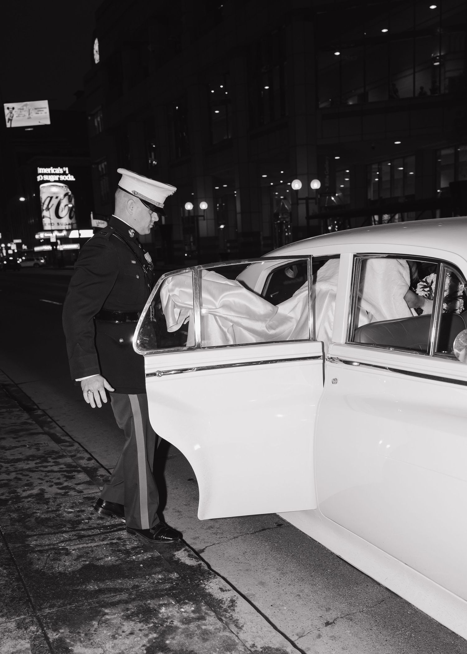 Black and white photo of Marine groom in dress uniform opening vintage car door for bride at night.