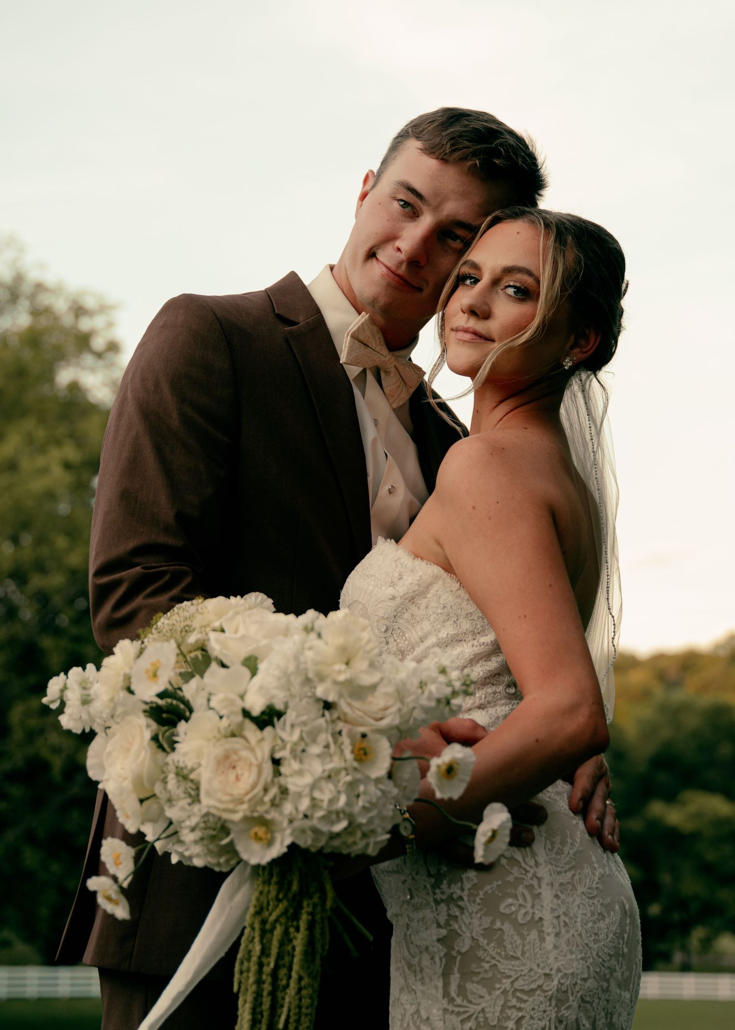 Bride in strapless lace gown and groom in brown suit embrace holding large white floral bouquet at dusk. Tennessee Wedding Photographer.