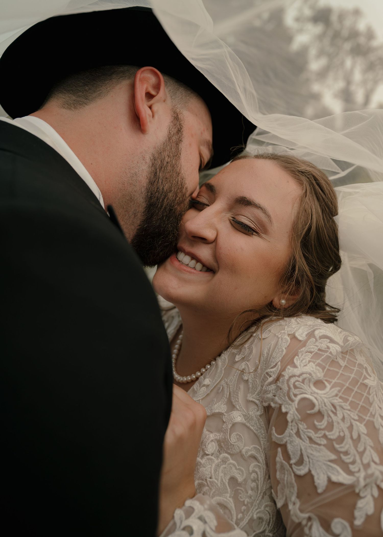 Groom in black hat tenderly nuzzling smiling bride in lace dress under flowing veil.