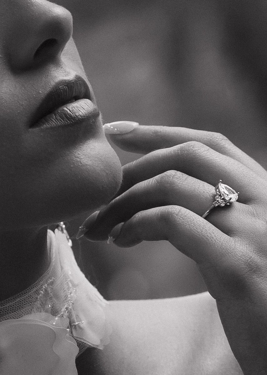 Black and white close-up of a bride wearing a pear-shaped diamond engagement ring, touching her chin.