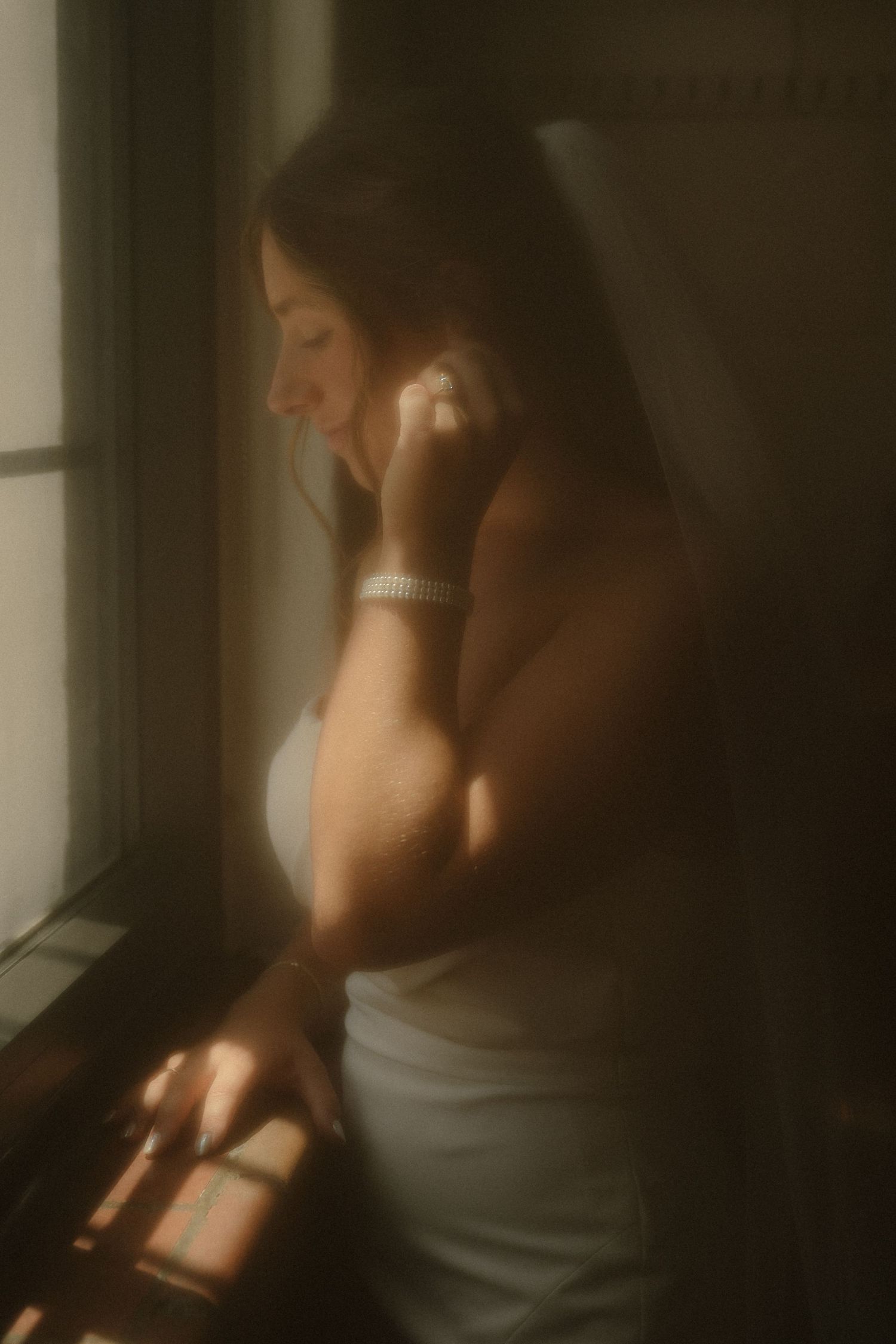 Bride in white dress standing by window in soft golden light, smiling while touching her face on wedding day.