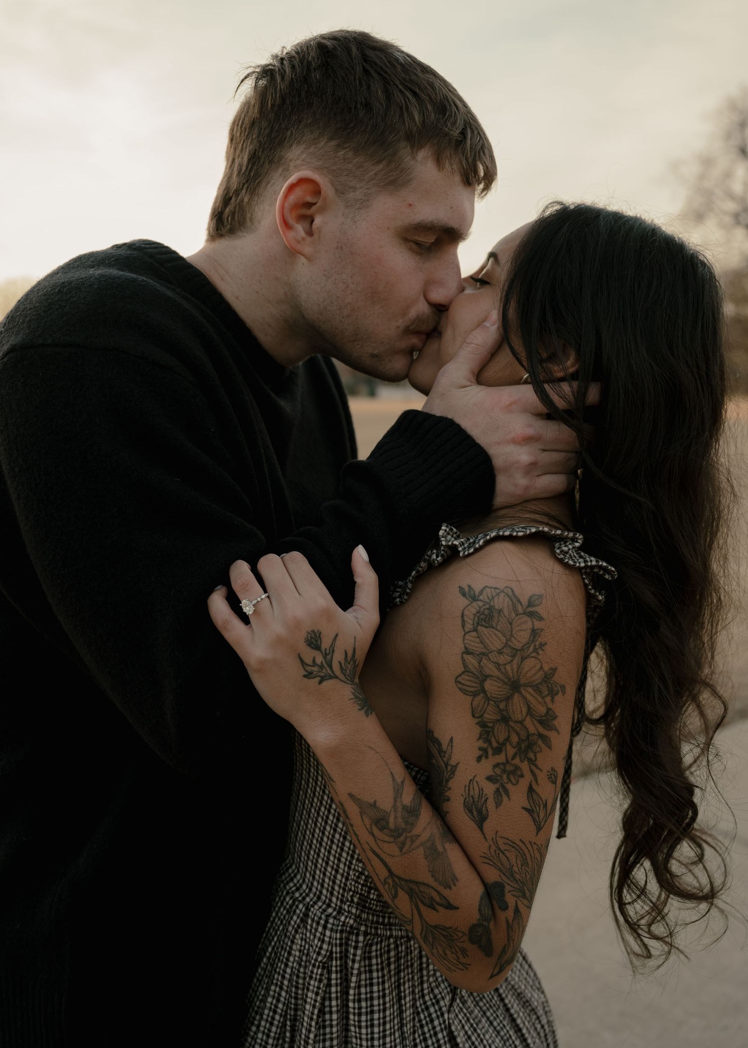 Tattooed couple sharing passionate kiss outdoors at dusk in close embrace.