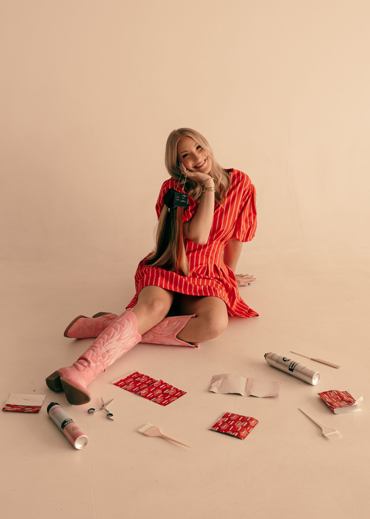 Smiling blonde woman in red striped dress and pink cowboy boots sitting on floor surrounded by wrapped gifts.