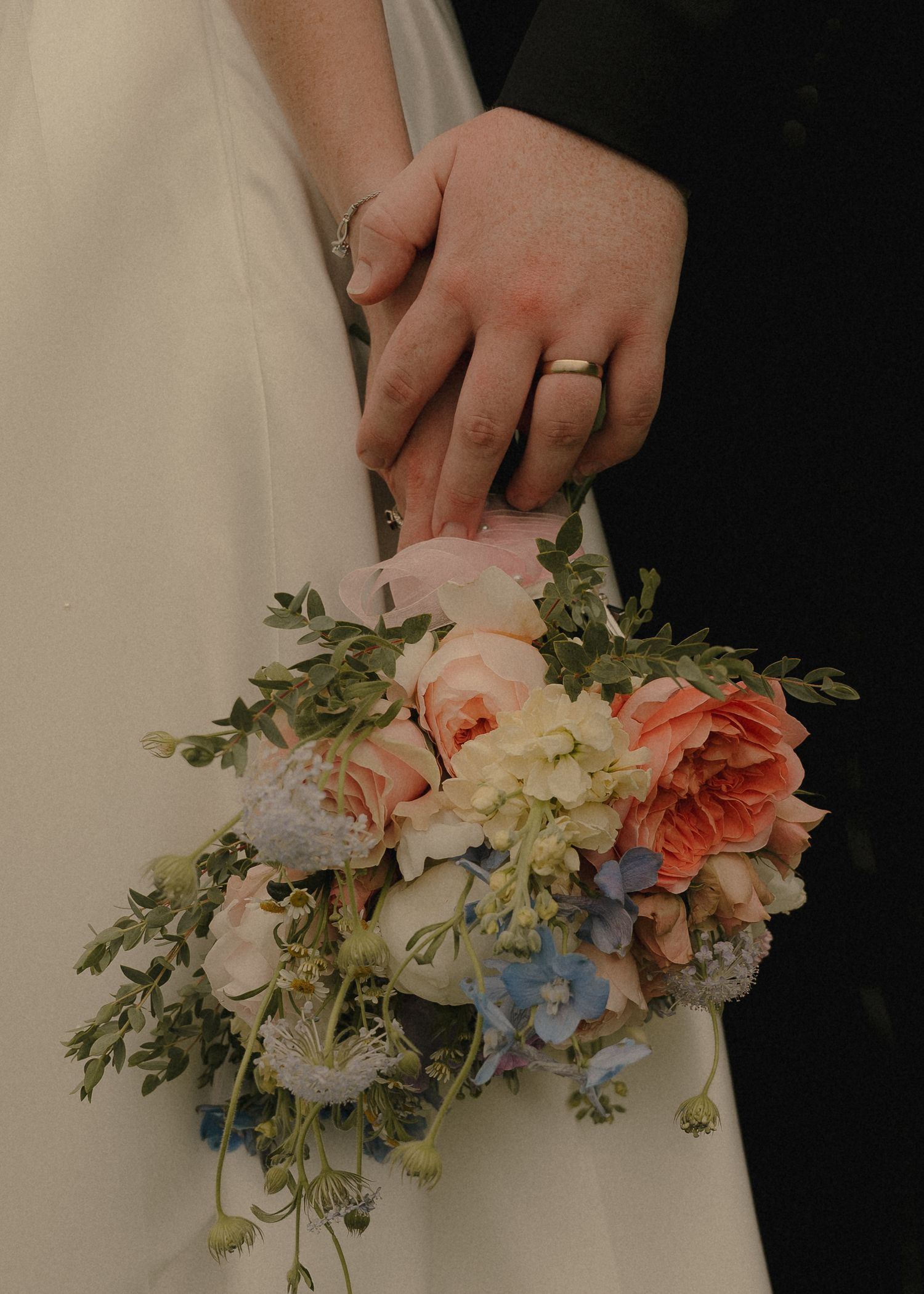 Close-up of bride and groom hands with wedding rings holding colorful bridal bouquet of roses.