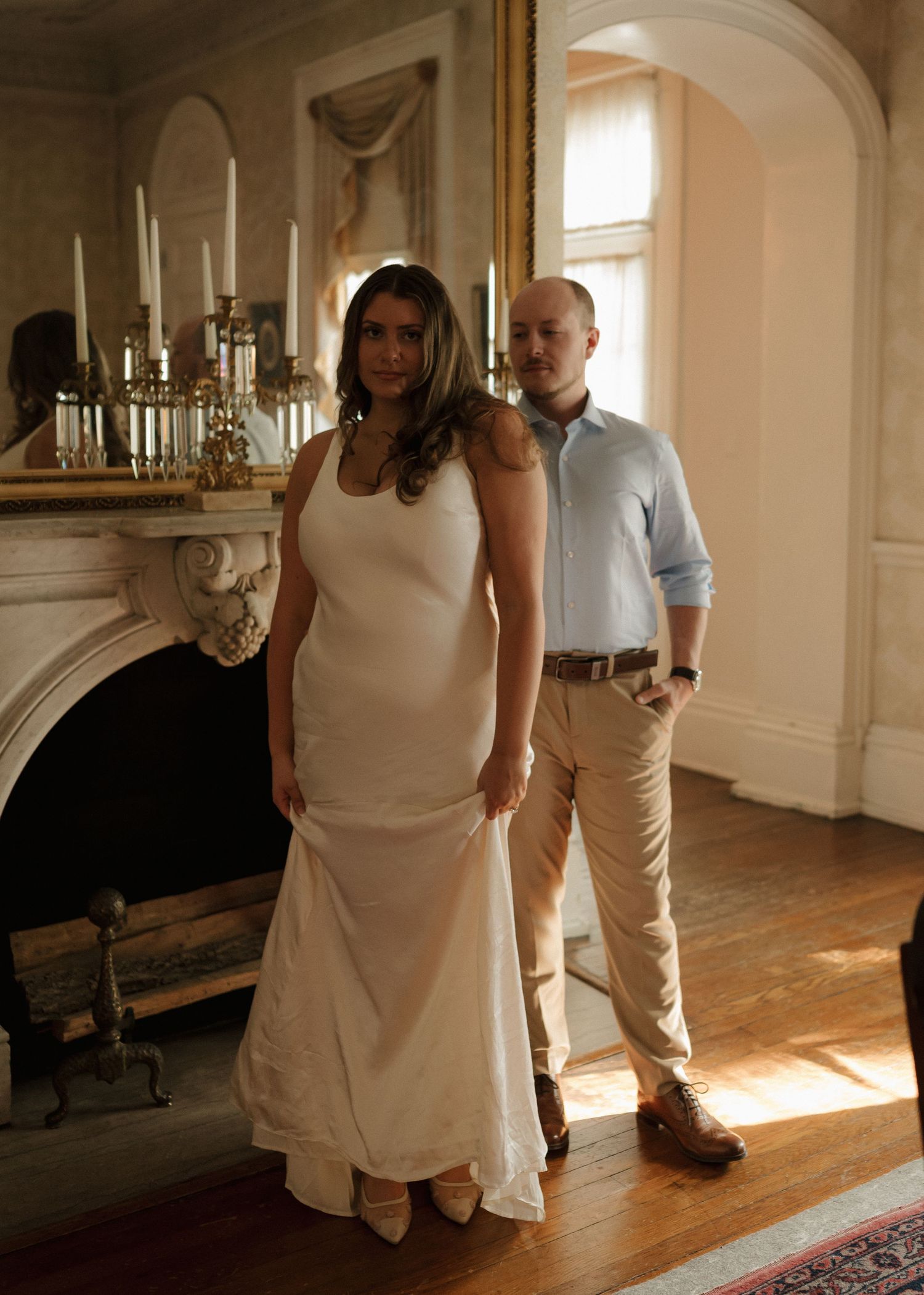 Bride in sleek white gown and groom in light blue shirt posing by a fireplace indoors.