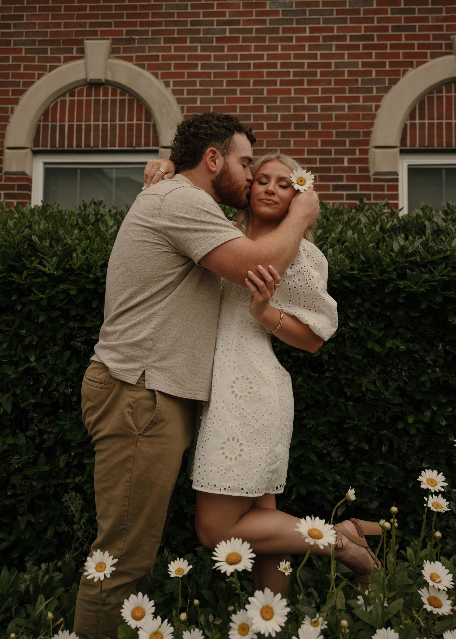 Couple sharing a romantic kiss among white daisies in front of a brick building with arched windows.