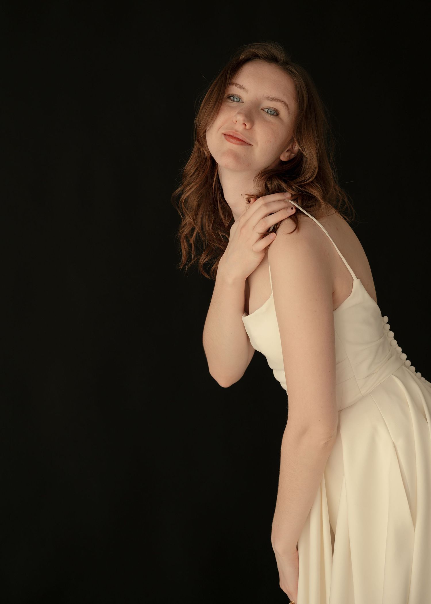 Young woman in white slip dress smiling softly against dark background in studio portrait.