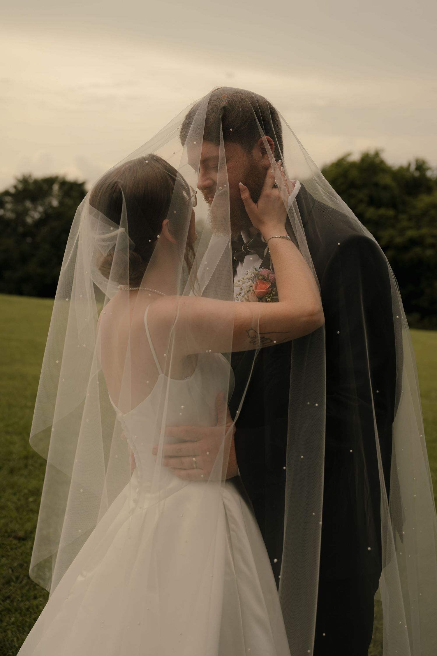 Bride and groom sharing intimate moment under pearl-adorned wedding veil in green field.